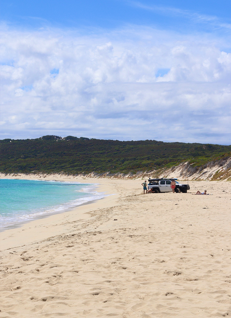 A sandy beach with a few people near a vehicle, overlooking the ocean with clear blue water and a green hill in the background, under a cloudy sky.