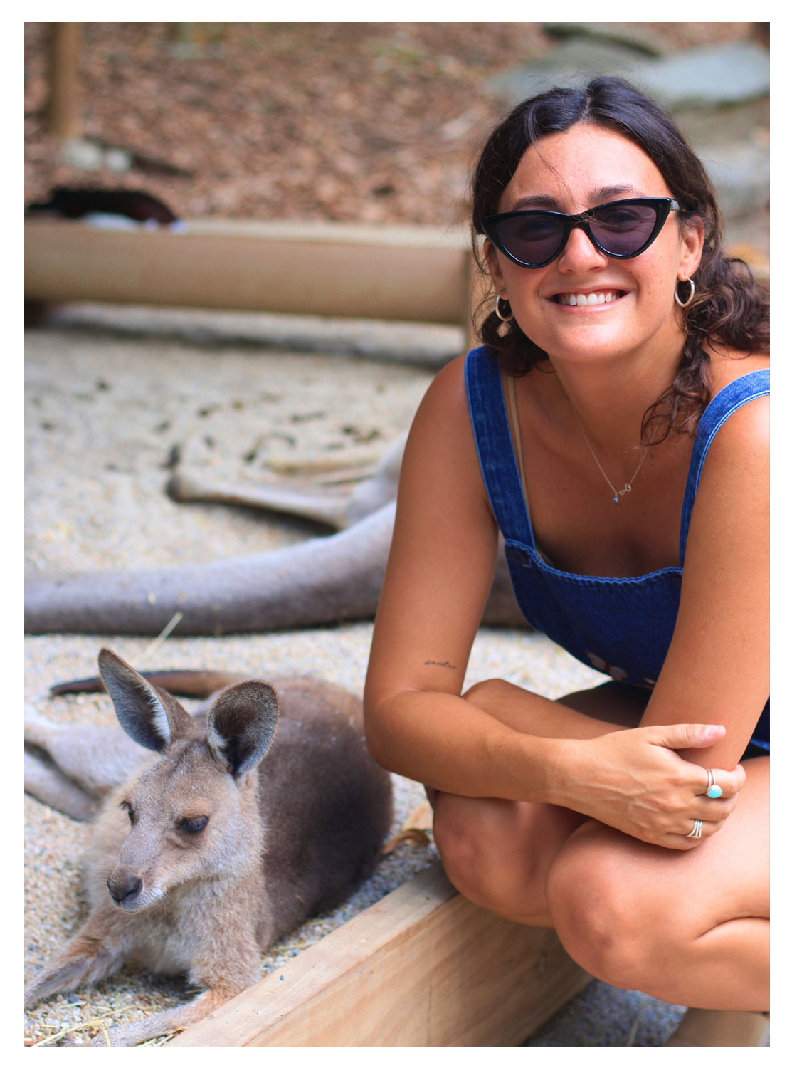 A woman with dark hair, wearing sunglasses and a blue dress, smiling while sitting next to a baby kangaroo on sandy ground.