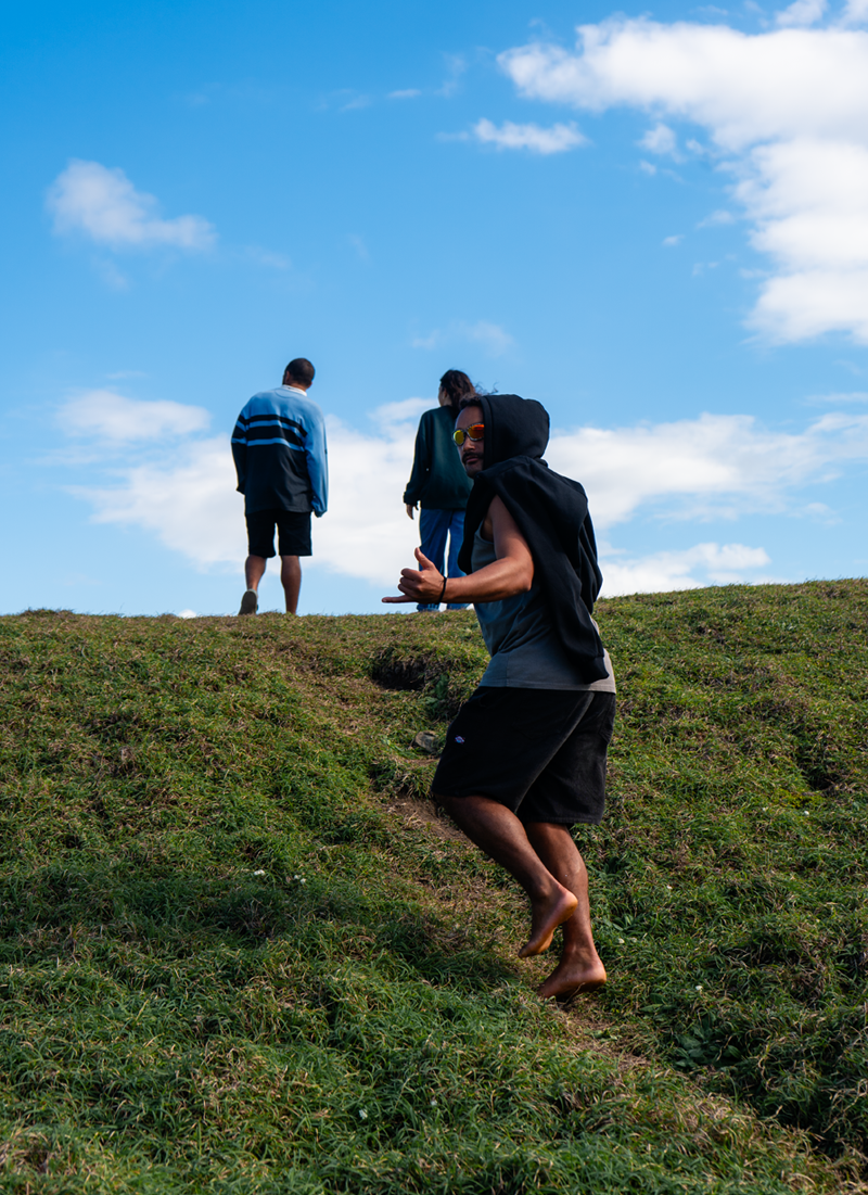 Three people outdoors on a grassy hill with a blue sky and clouds, one climbing uphill and two standing at the top.