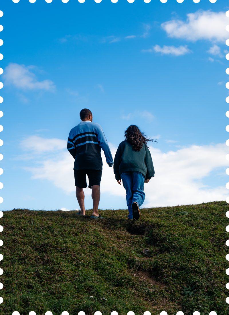 Two people walking up a grassy hill, facing away from the camera, with a blue sky and clouds overhead.