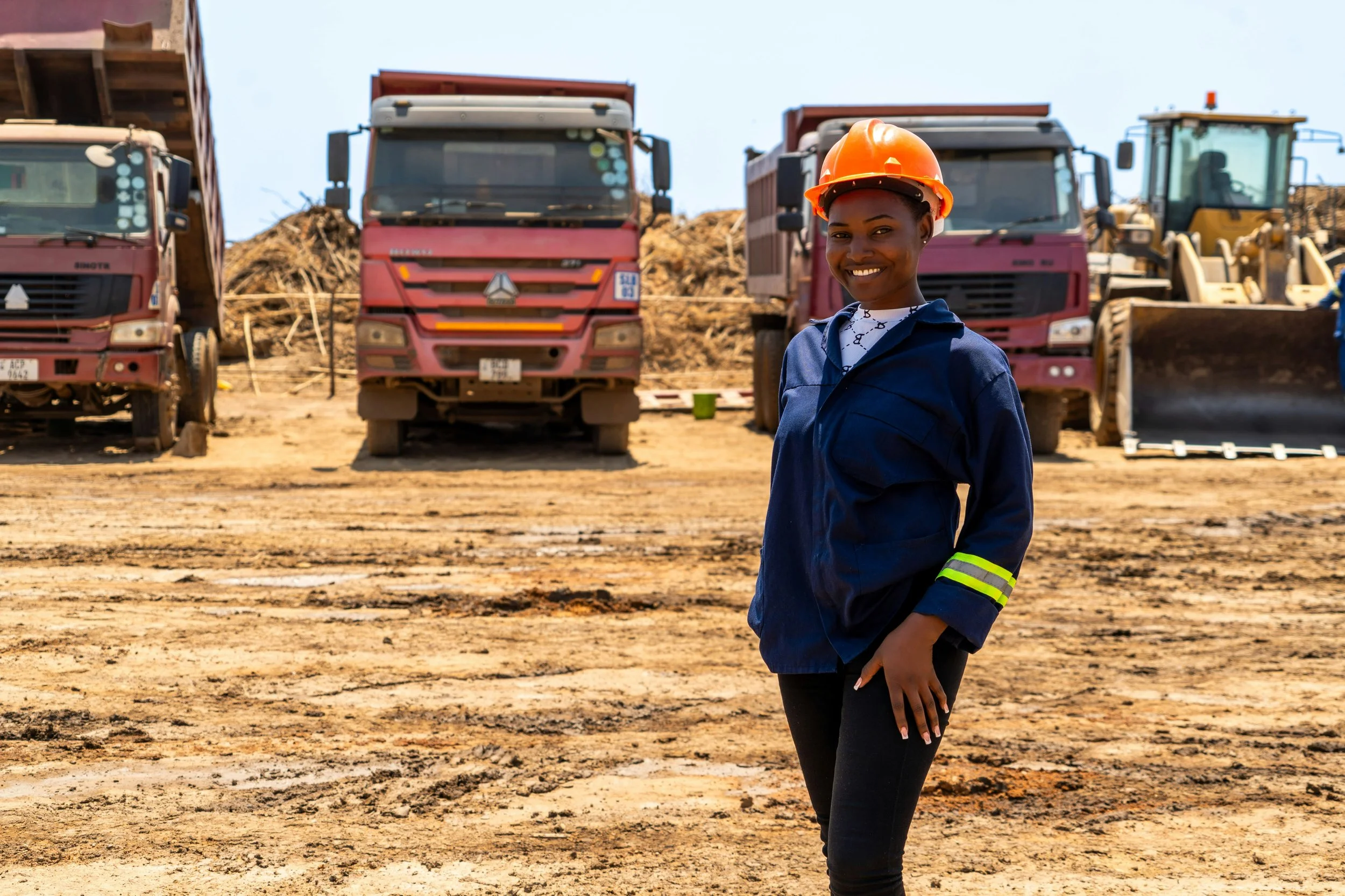 A woman smiling wearing an orange safety helmet and dark blue jacket with reflective stripes on the sleeves, standing in a construction or excavation site with large trucks and construction equipment in the background.