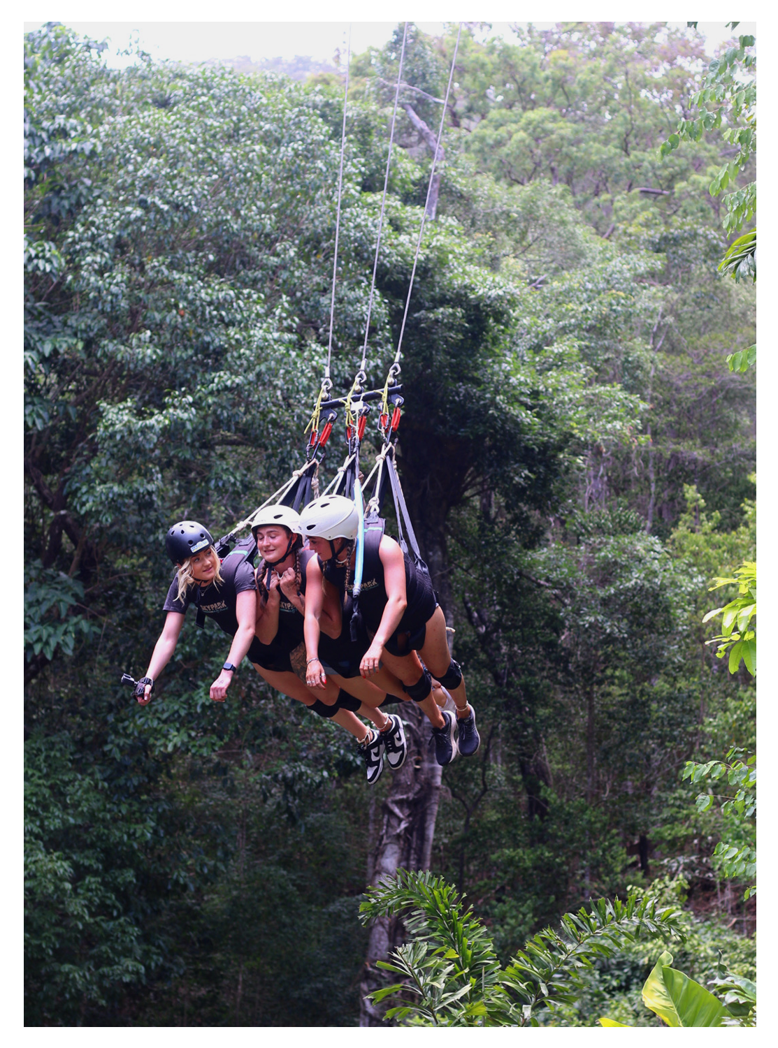 Three women in helmets and harnesses hang from a zip line in a lush green forest.