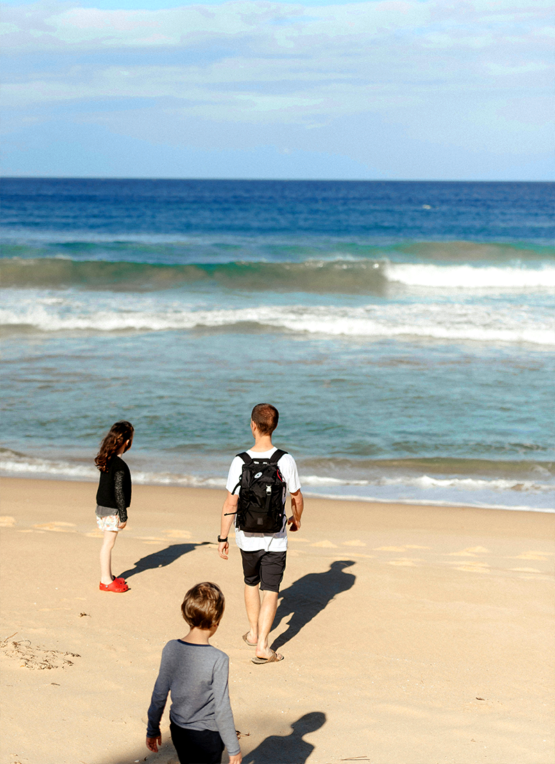 Three people, two children and one adult, walking on a sandy beach towards the ocean.