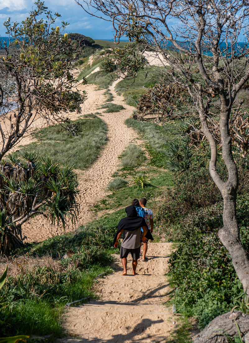 Three people walking along a sandy trail on a coastal hillside, surrounded by sparse trees and green vegetation, with sand dunes and the ocean in the background on a partly cloudy day.