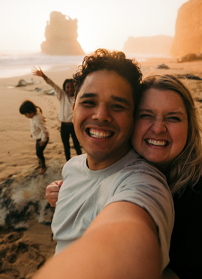 A group of four friends taking a selfie on the beach during sunset, with cliffs in the background, smiling and enjoying their time.