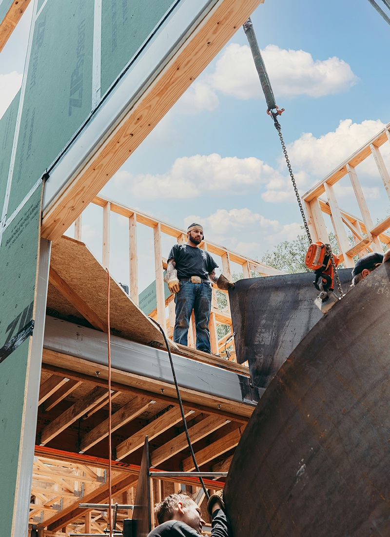 A construction worker standing on a wooden framework at a building site, operating a crane to move a large steel component, with a blue sky and white clouds in the background.