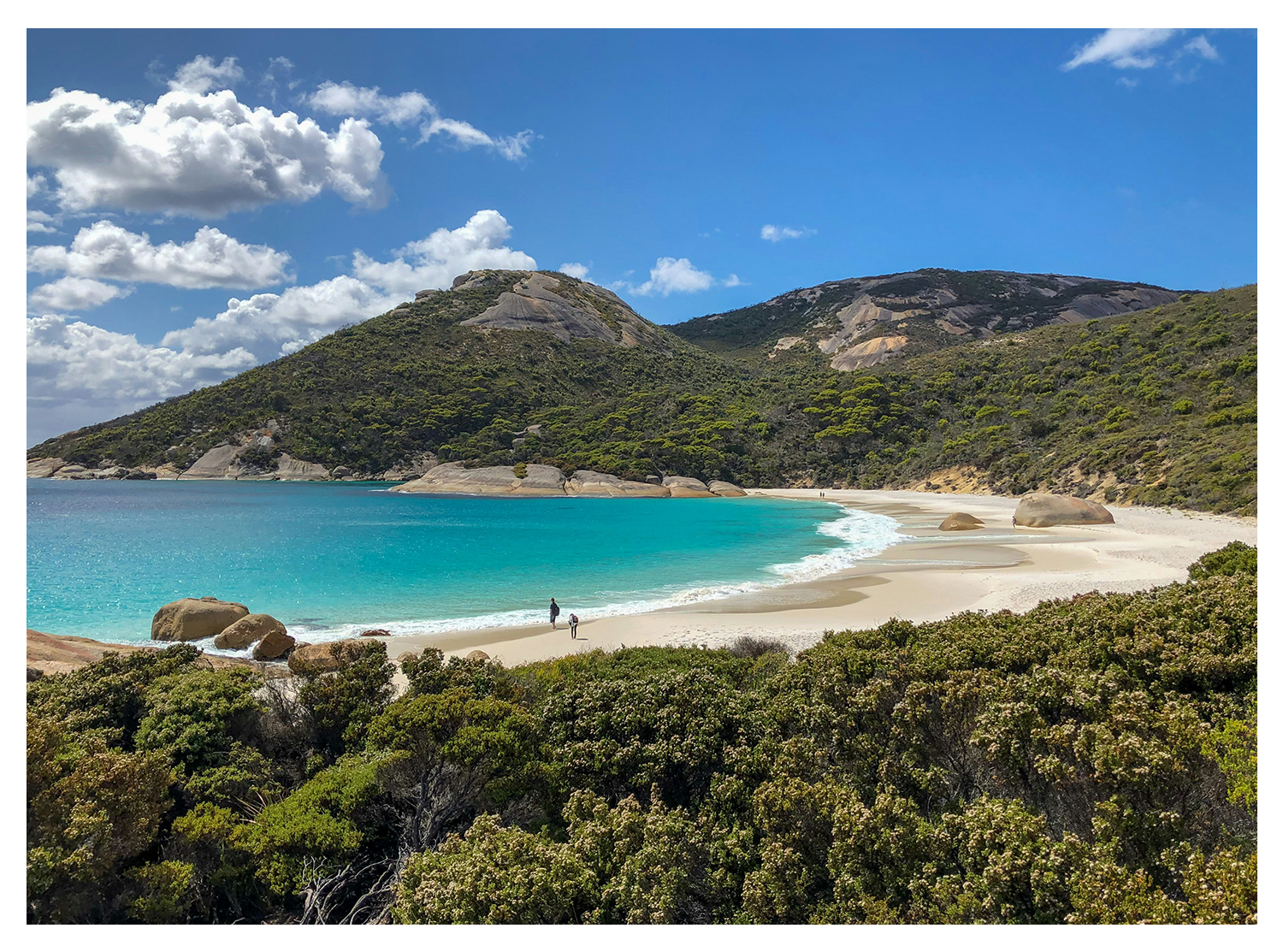 A scenic beach with white sand, turquoise water, large rocks, and green hills under a partly cloudy sky, with two people walking along the shore.
