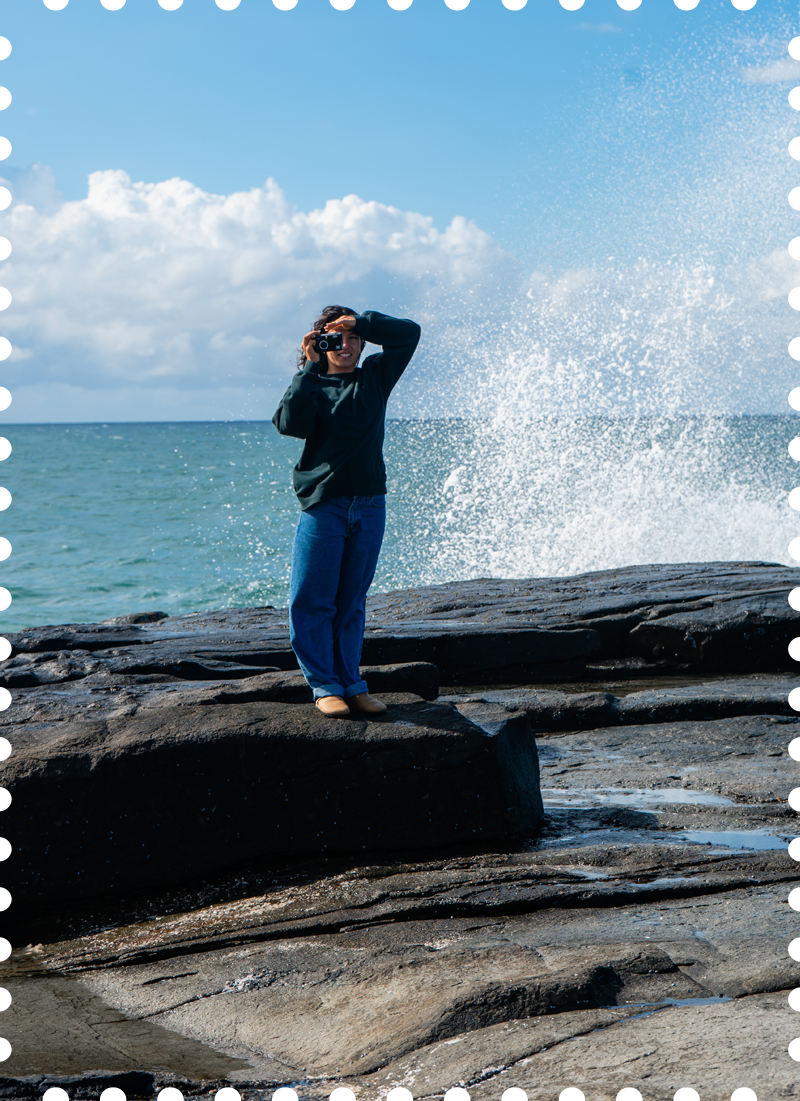 A person standing on rocks by the ocean, holding a camera and looking through it while waves splash behind them under a partly cloudy sky.