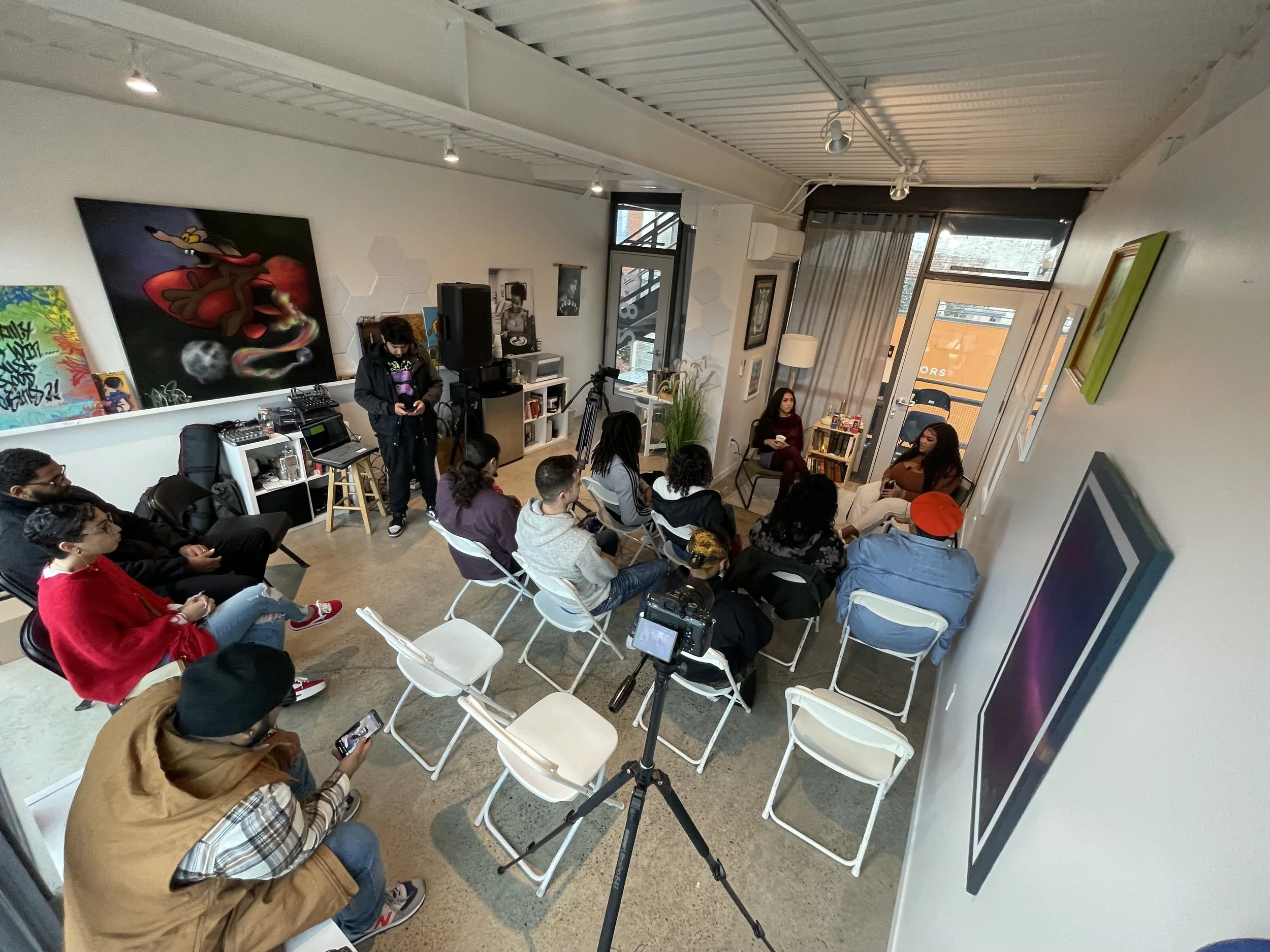 People gathered in a room attending a presentation or workshop, sitting on chairs and listening. There are artworks on the walls and a tripod with camera equipment in the room.