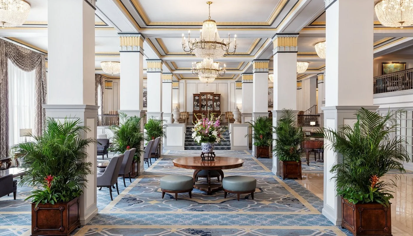 Elegant hotel lobby with high ceilings, chandeliers, potted plants, and a central round table with floral arrangement.