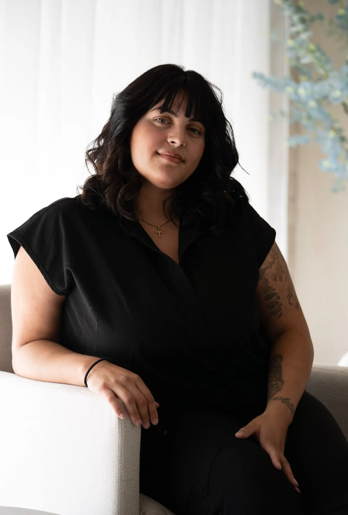 A woman with black hair and tattoos on her right arm sitting on a cream-colored couch in front of a white curtain, wearing a black top and a cross necklace.