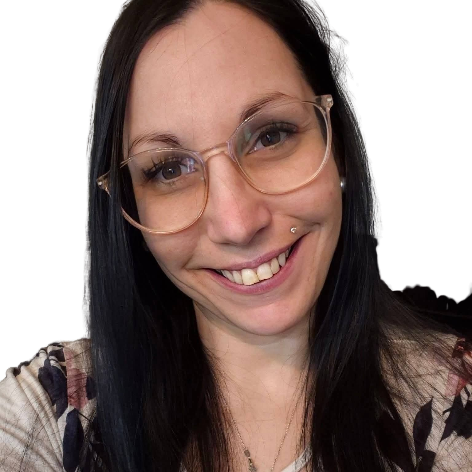 Une femme souriante avec des lunettes transparentes, des cheveux longs et noirs, portant une robe à motifs floraux, et un collier pendentif.