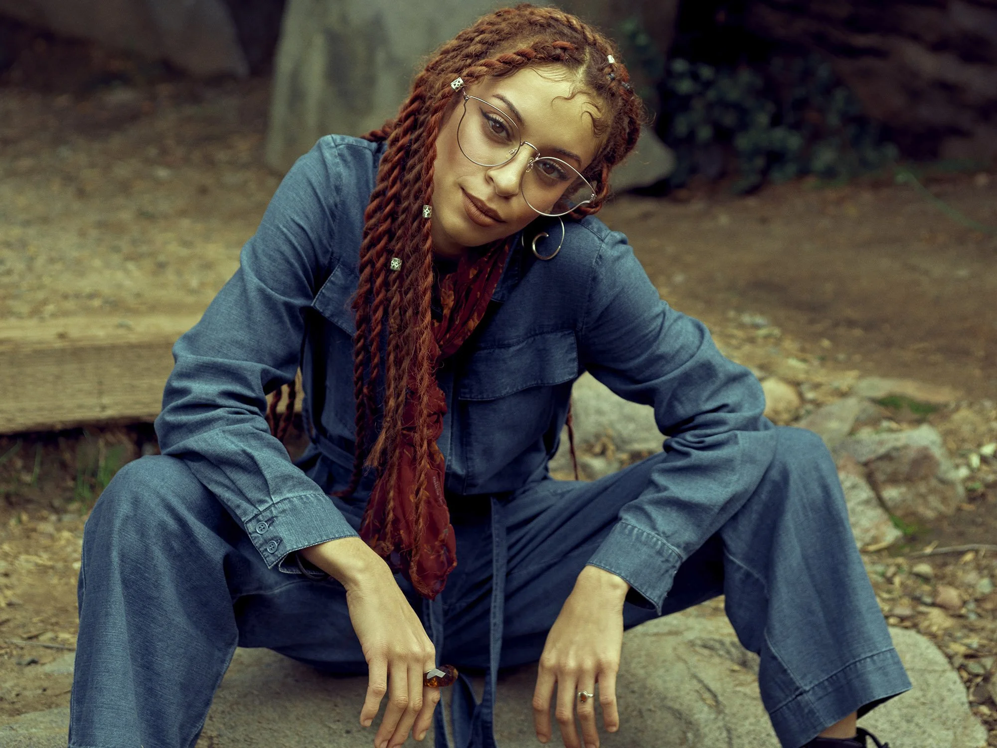 A woman with red dreadlocks and glasses, dressed in blue denim, sitting outdoors with a natural background, looking at the camera.