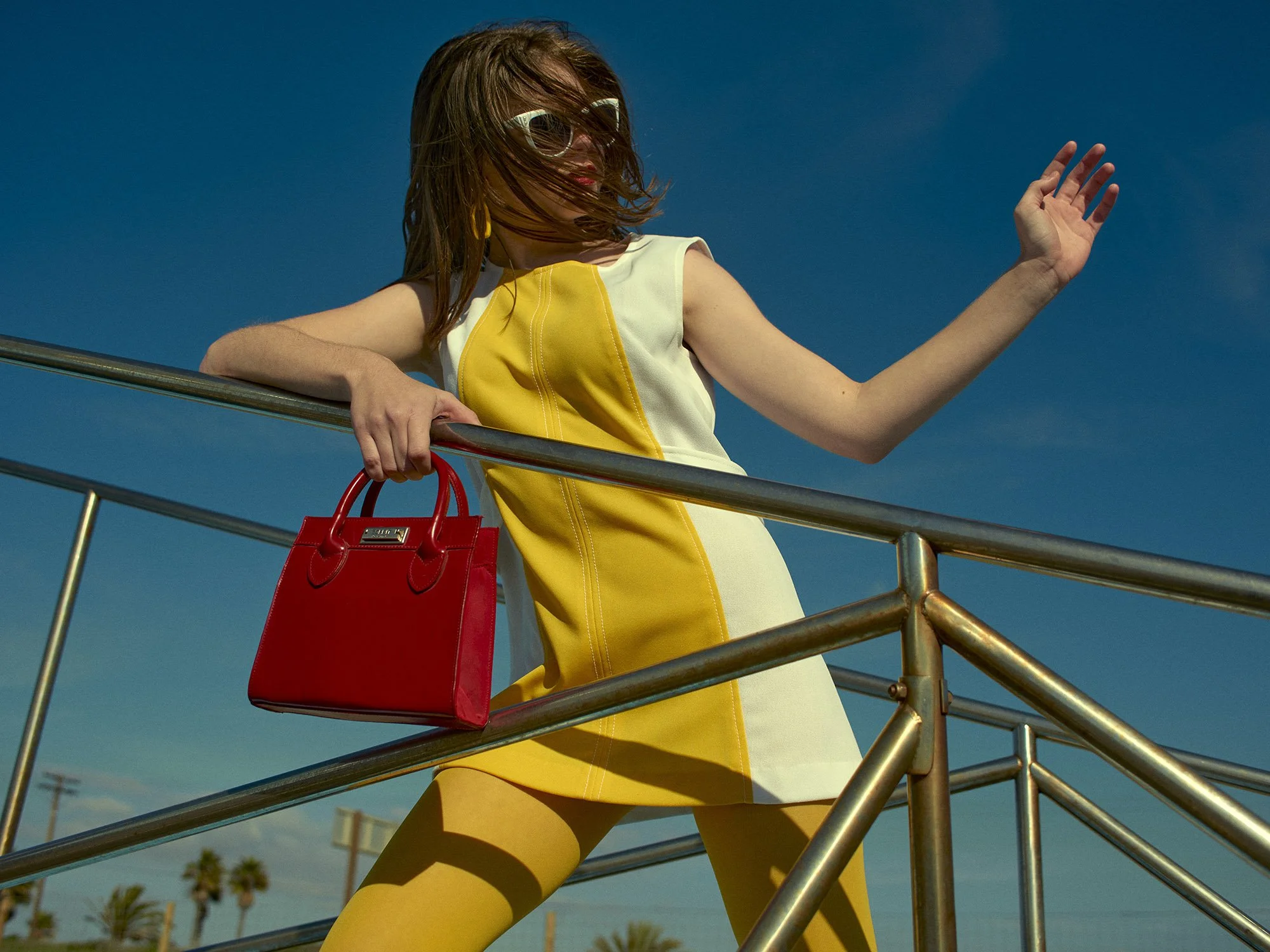 A woman wearing sunglasses, a yellow and white dress, and yellow tights, holding a red handbag, stands on a metal railing against a bright blue sky with palm trees in the background.