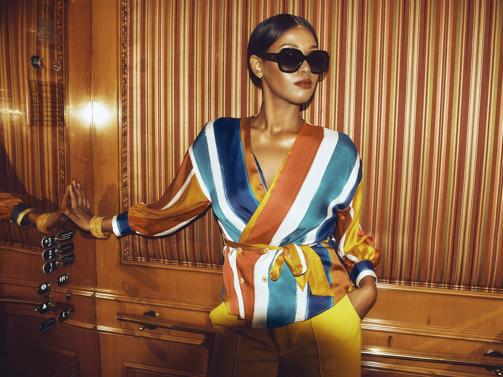 A woman with dark hair wearing large black sunglasses, a colorful striped silk blouse, and yellow pants standing in an elevator with wood paneling and control panel.