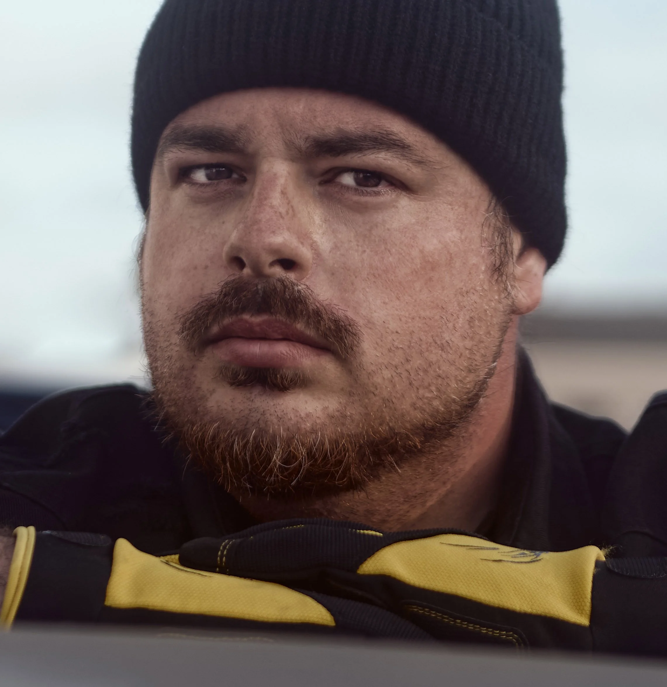 Close-up of a person's face wearing a black beanie and a yellow jacket. Only the upper part of the face is visible, including eyes, eyebrows, nose, and part of facial hair. The background is blurred and appears to be outdoors.