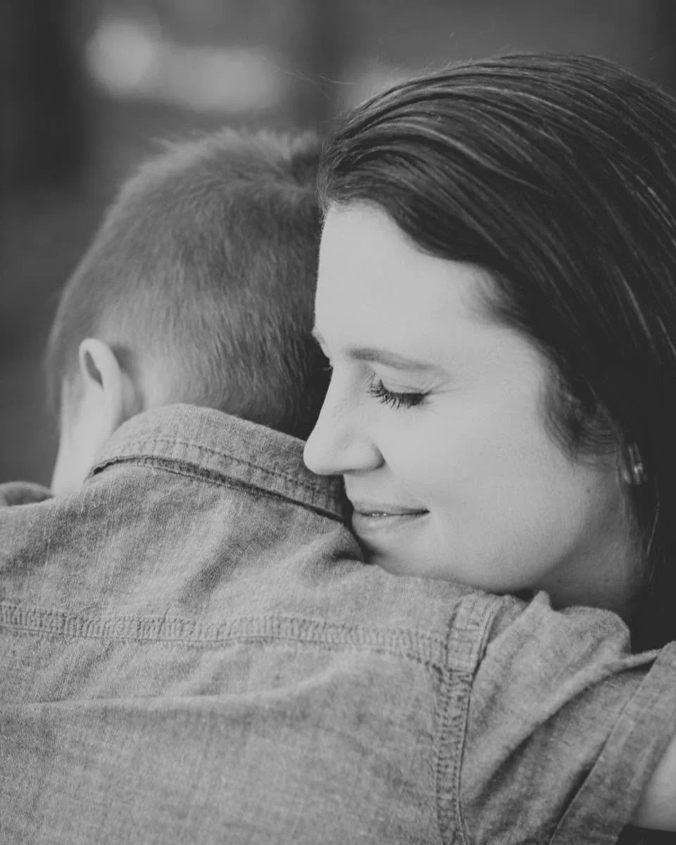 A woman with dark hair and closed eyes embraces a young boy with short hair, both shown from the side, in a black and white photograph.