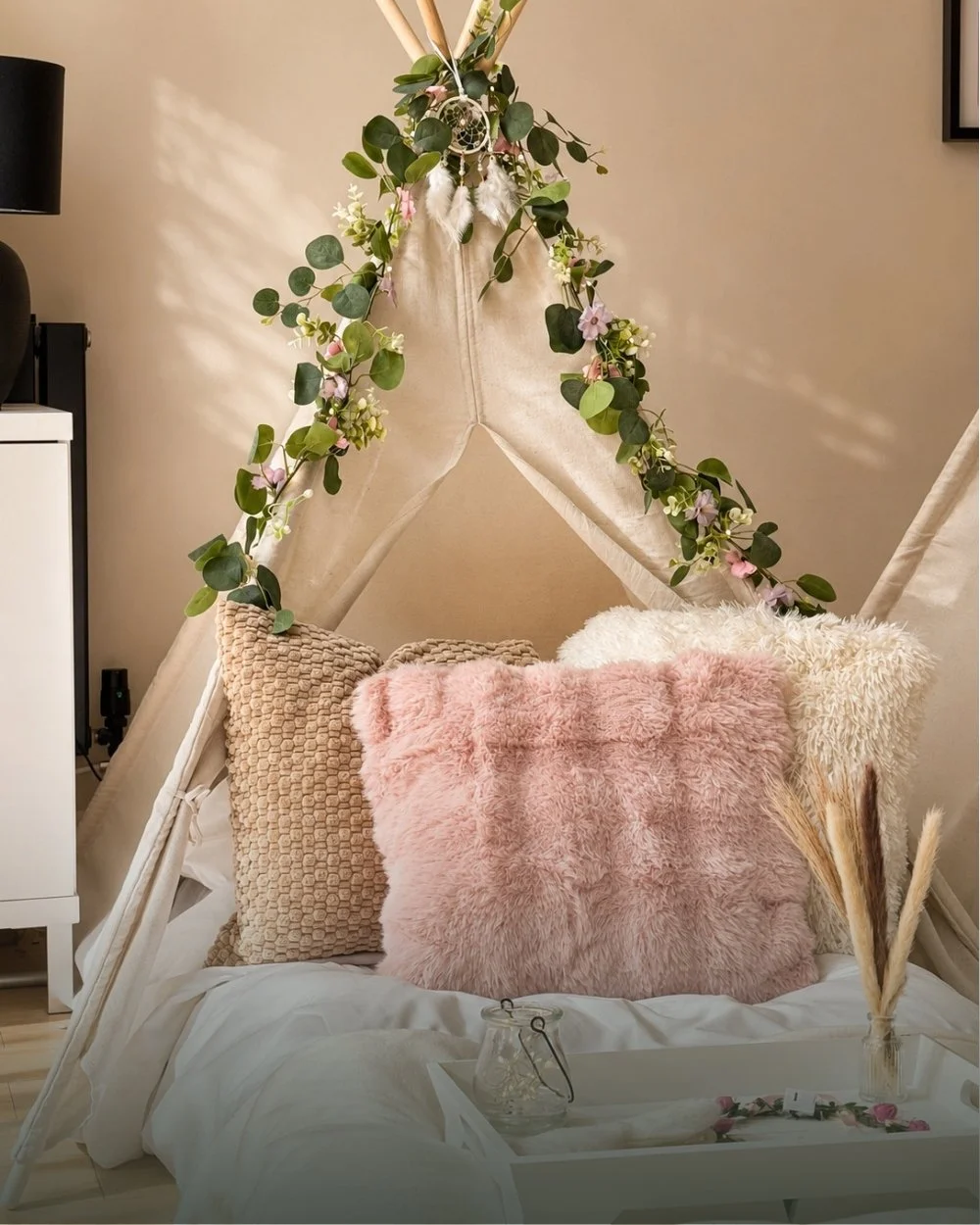 A boho styled teepee with a floral garland, dreamcatcher, pampas grass and soft neutral cushions