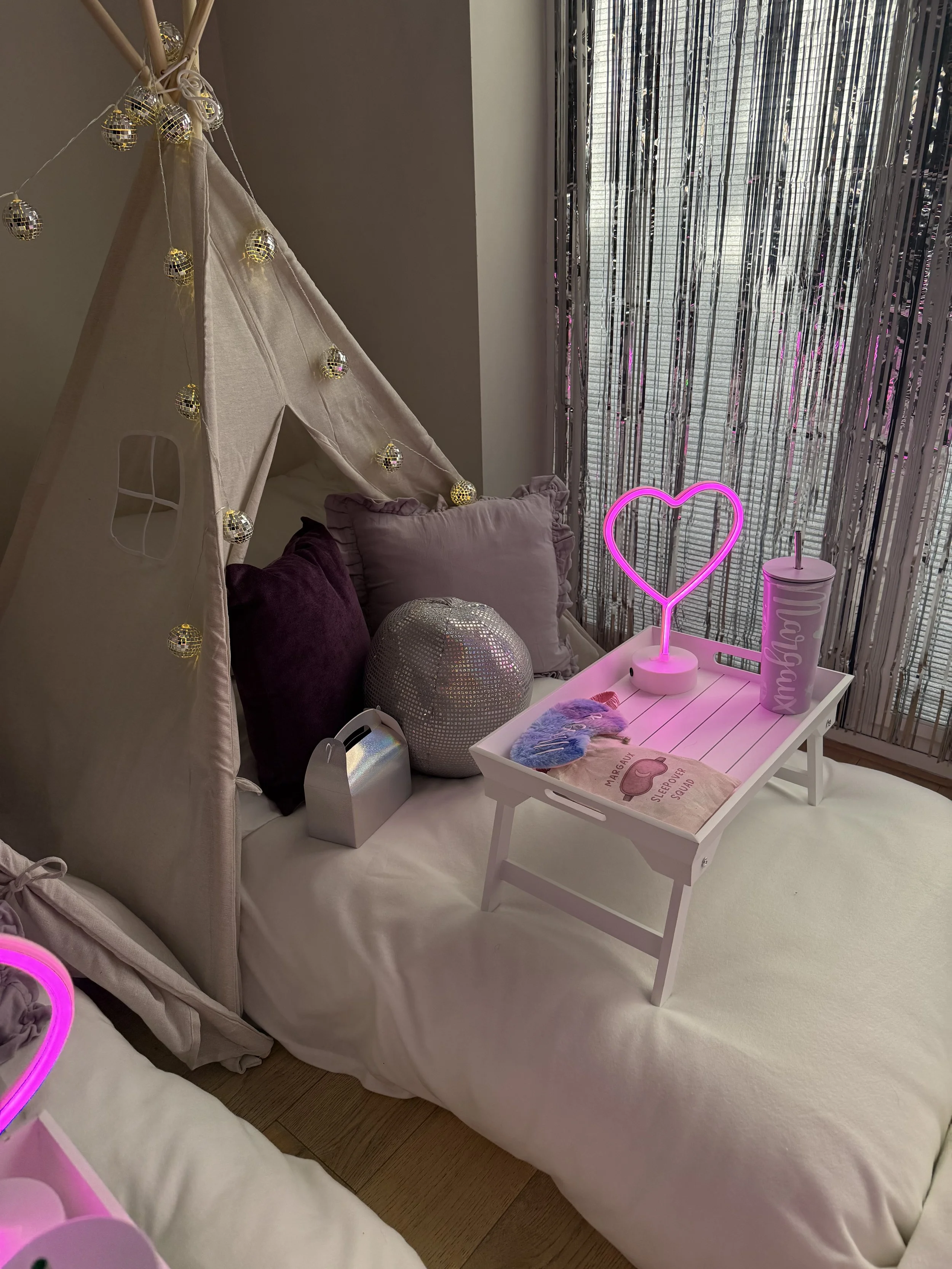 Glitterball Disco Dreams teepee sleepover party by Sweet Dreams Events showing a cream teepee with disco ball fairy lights, neon pink heart lamp silver tinsel backdrop, personalised cup and styled breakfast tray set up for a girls party in Surrey