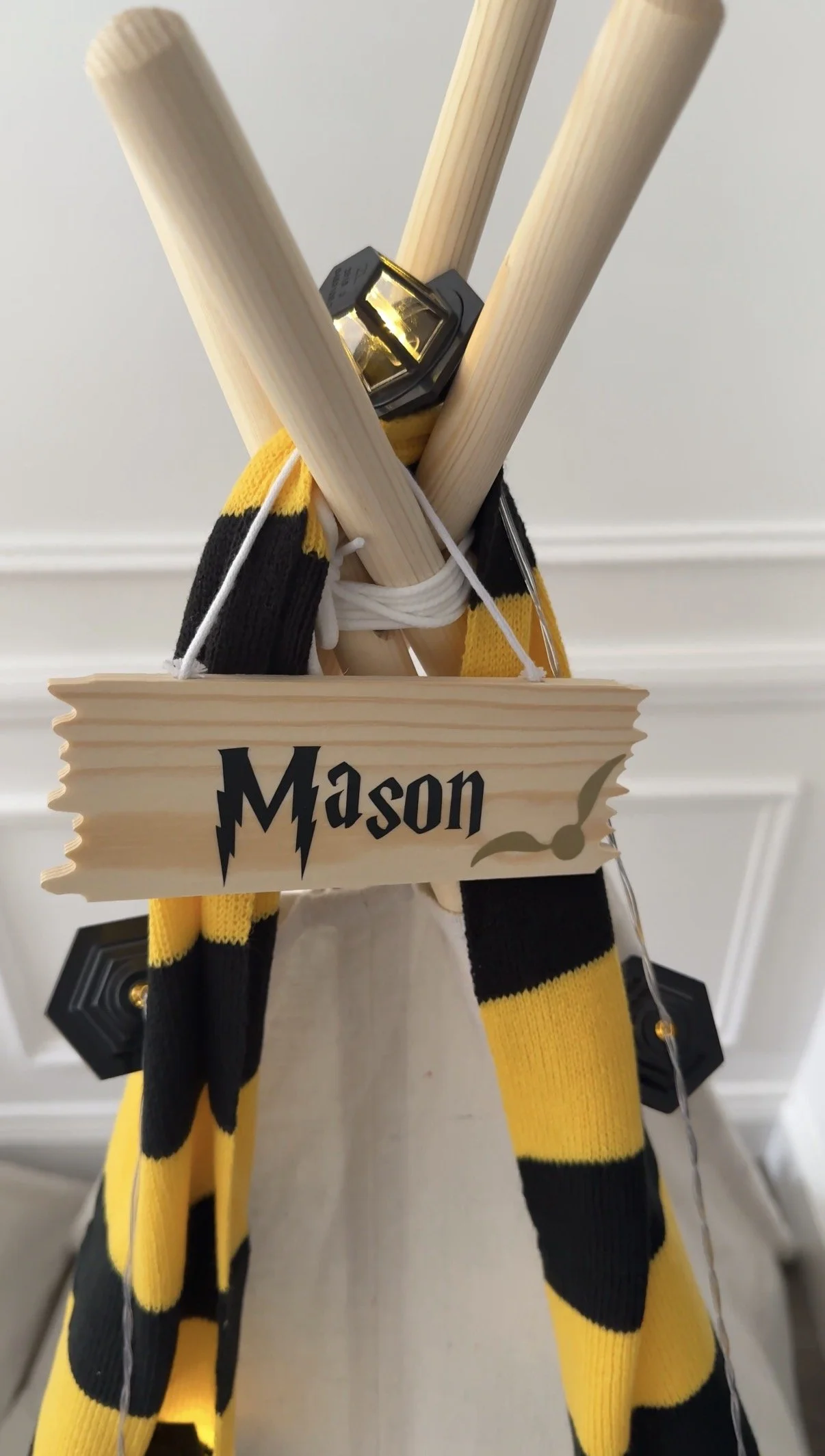 Personalised wooden name sign hanging from a wizard themed teepee with a yellow and black house scarf and glowing lights