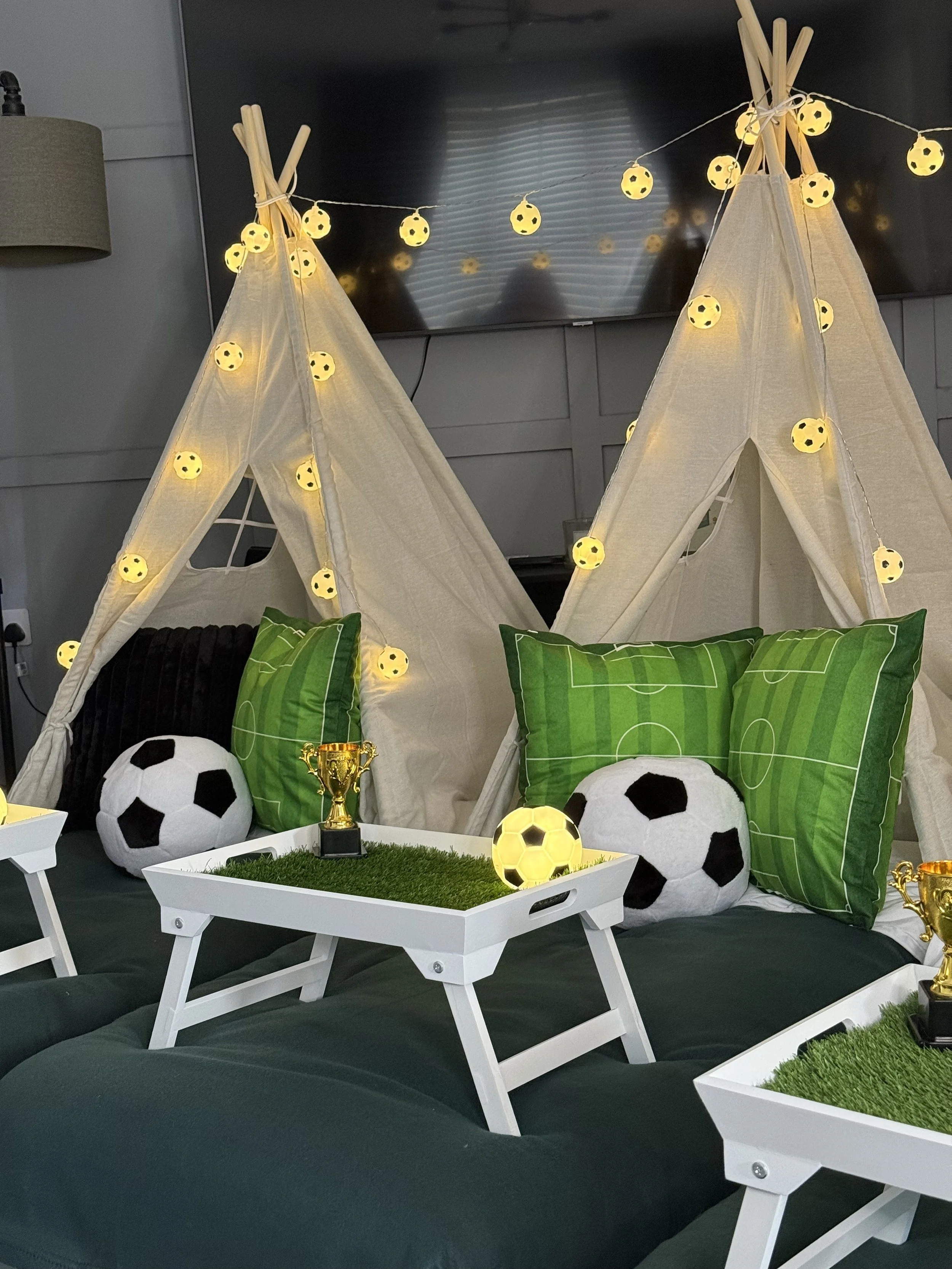 Living room decor themed around soccer with draped teepees decorated with string lights shaped like soccer balls, green pillows with soccer field graphics, plush soccer ball cushions, a white tray with a small trophy, and a black couch.