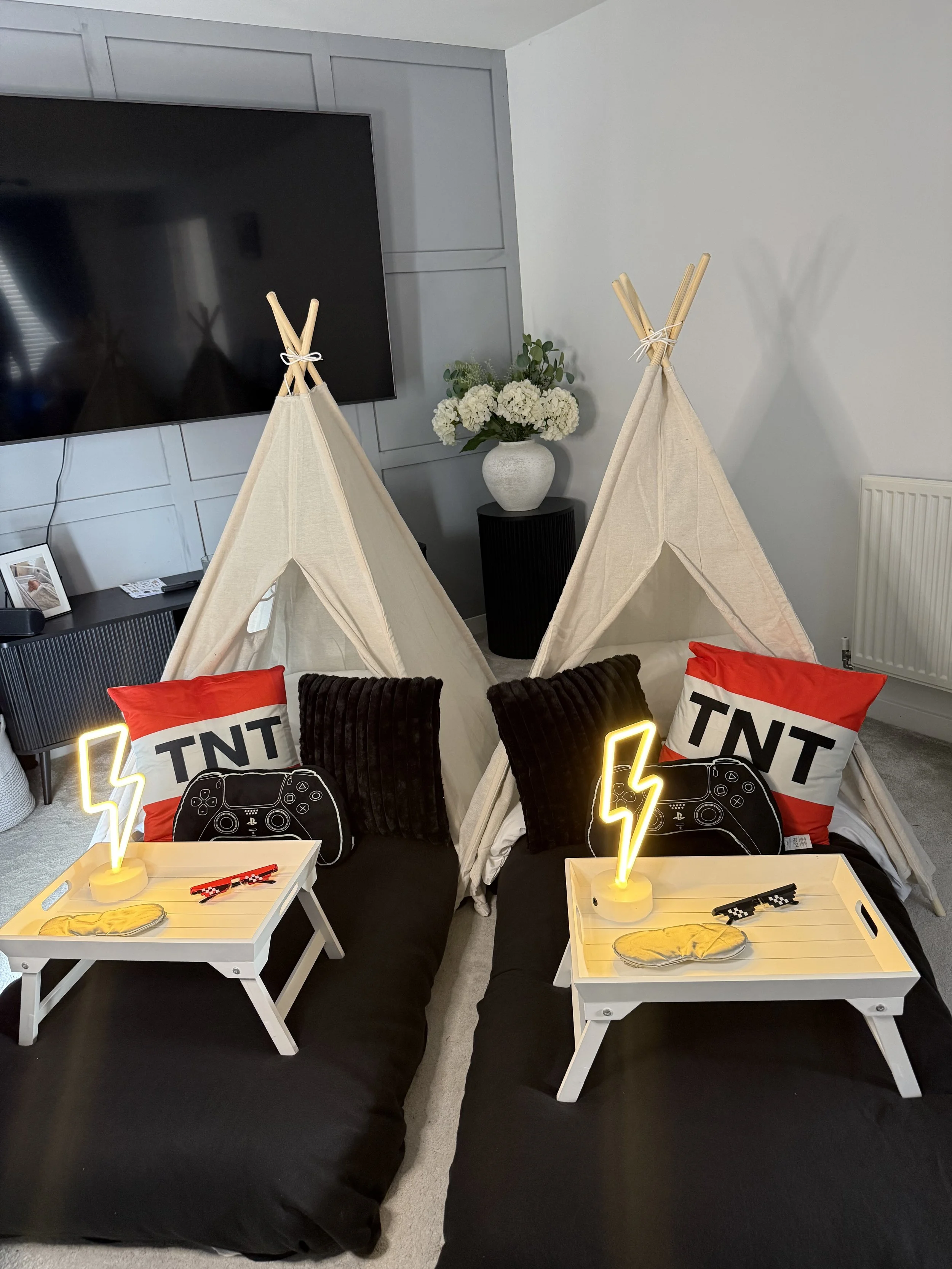 Living room setup with two small tepees, two black pillows, and two white trays on the floor. Each tray has a yellow neon lightning bolt decoration, a game controller graphic, a pair of yellow slippers, and a red and white TNT pillow. In the backgrou