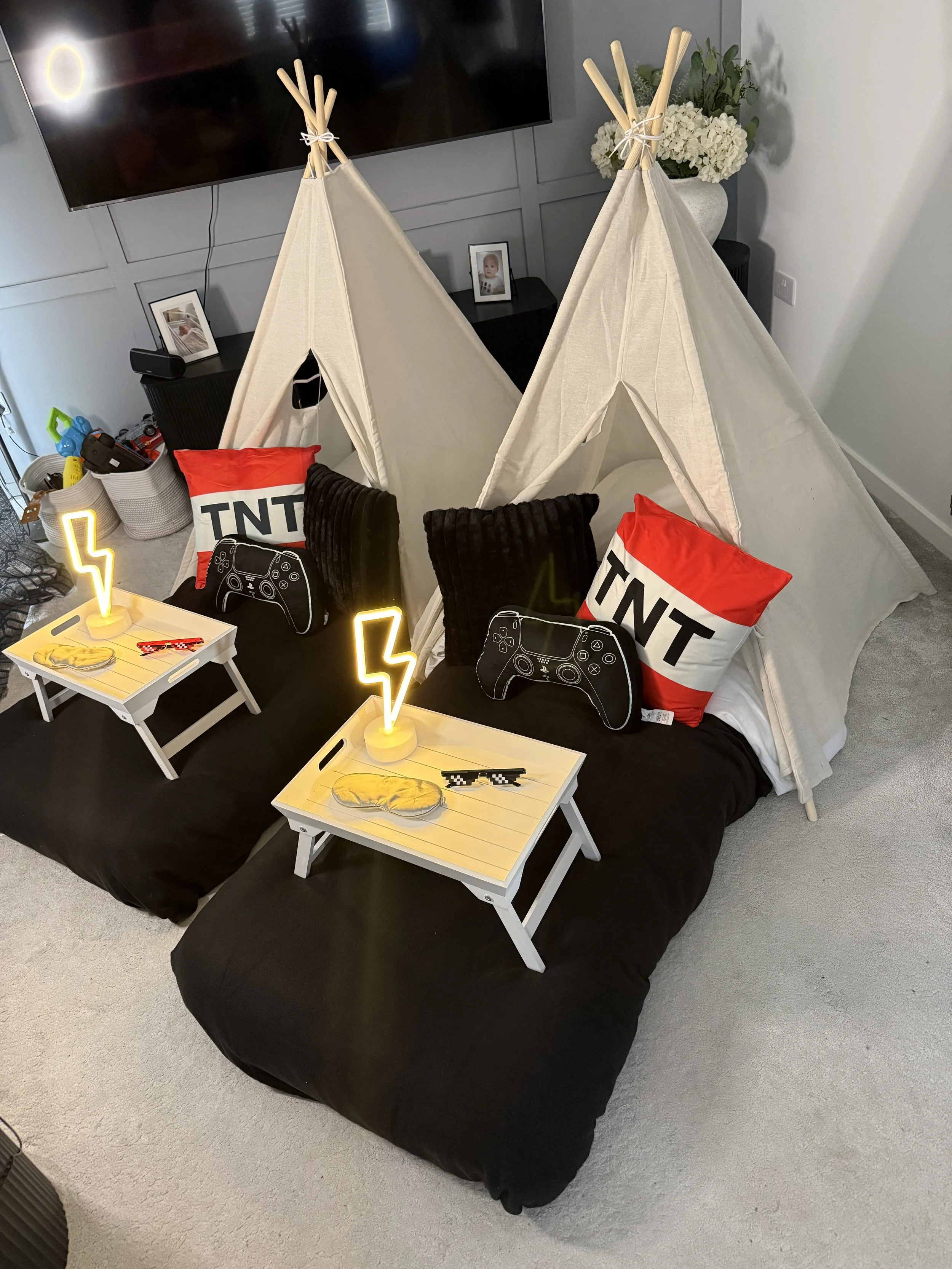 Two miniature beds with black bedding, decorated with pillows with 'TNT' and gaming controller designs, are set up on a large couch or bed. Each has small white trays with neon yellow lightning bolt lamps and metallic decorative rocks. The bed area i