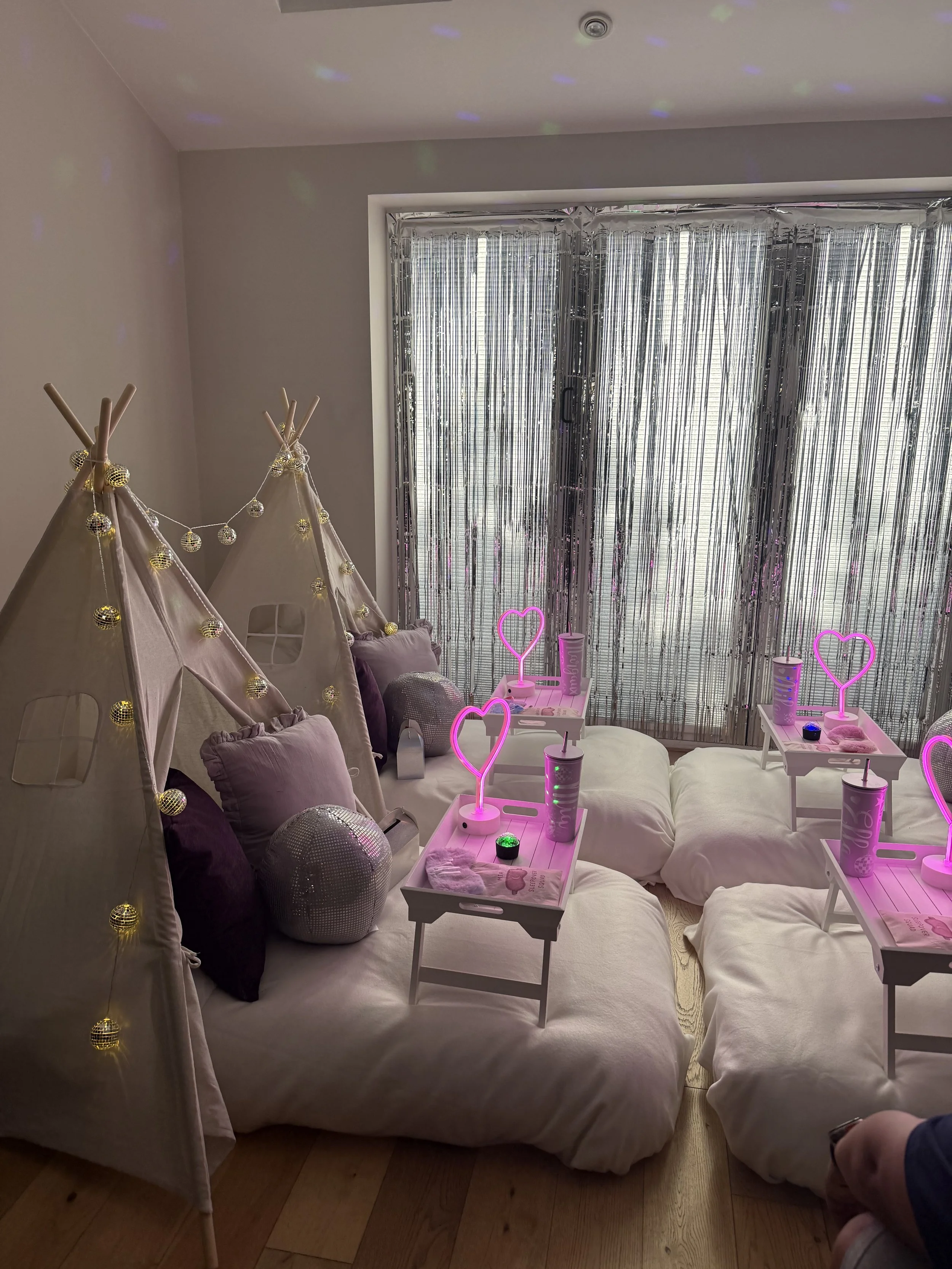 A cozy indoor space decorated with small tents, string lights, purple pillows, and parasol-shaped lamps, with a window covered by metallic silver curtains.