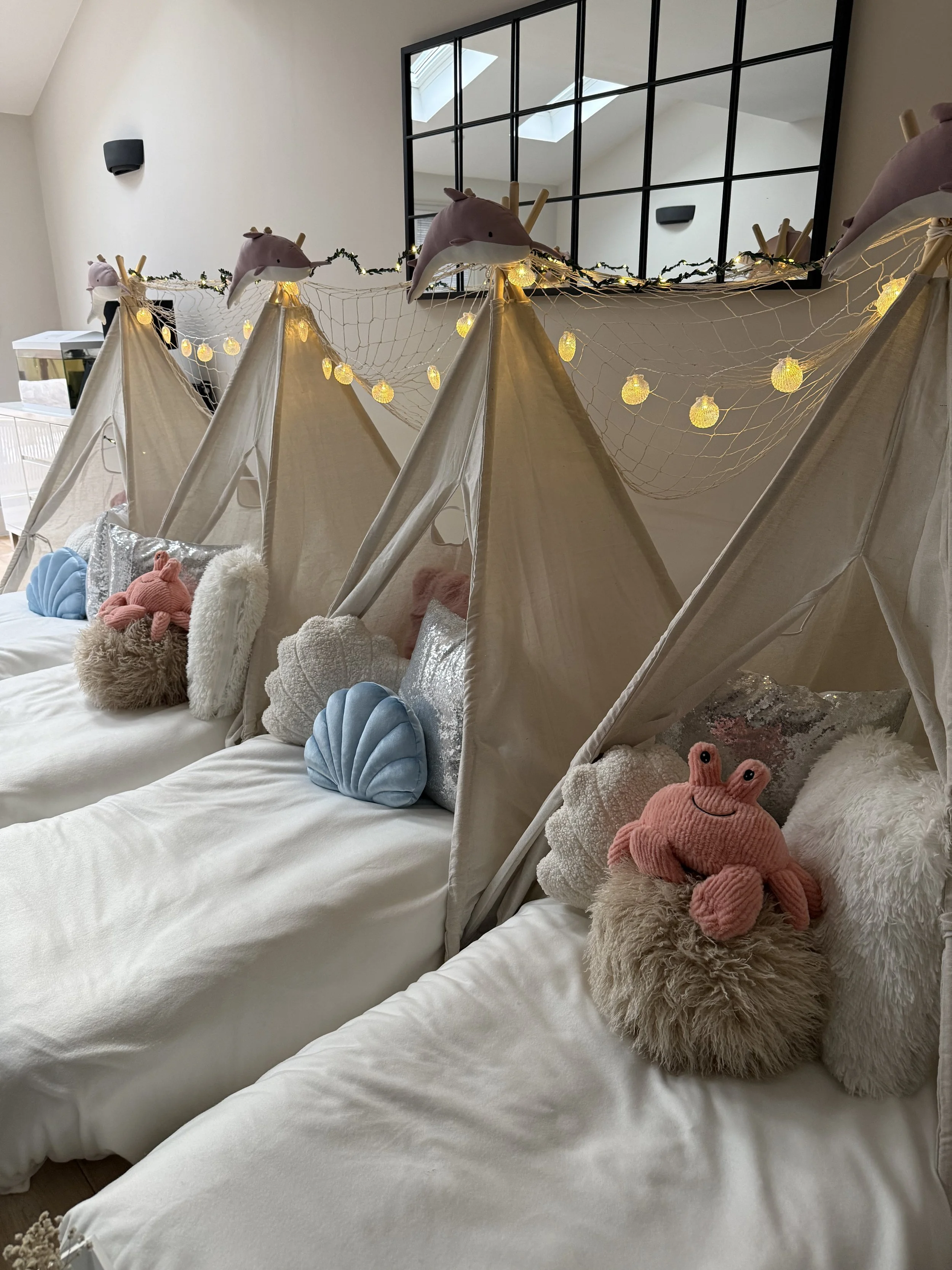 Mermaid Lagoon themed sleepover party with four white teepees, pink dolphin toppers, seashell fairy lights, blue velvet shell cushions, and a plush pink crab keepsake by Sweet Dreams Events