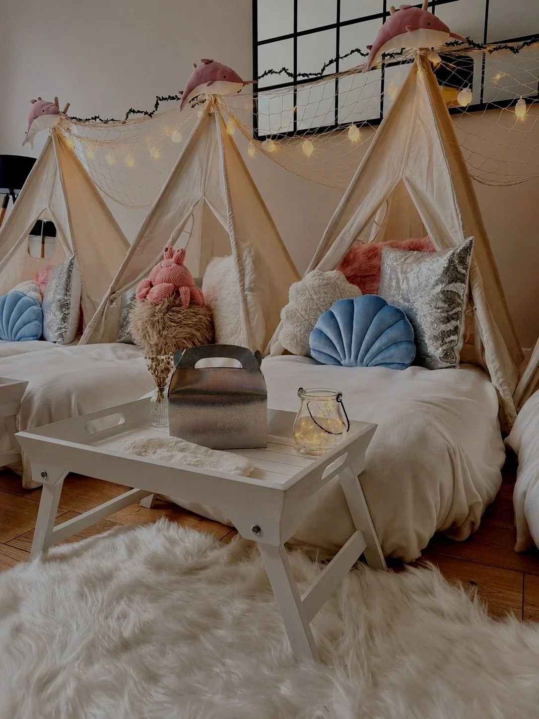 A cozy bedroom featuring a bed with multiple decorative pillows, including plush and sequined ones, and a canopy made of sheer fabric with string lights. The room has a soft, warm ambiance with a nightstand and minimal decor.