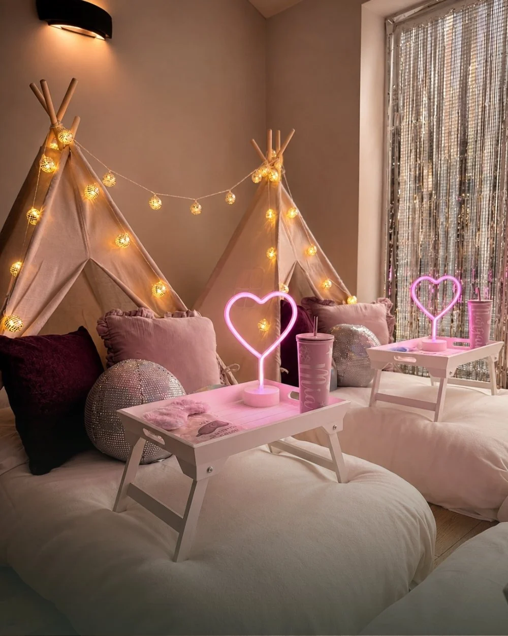 Two fairy lit teepees styled for a disco sleepover with pink neon hearts, disco balls and personalised trays