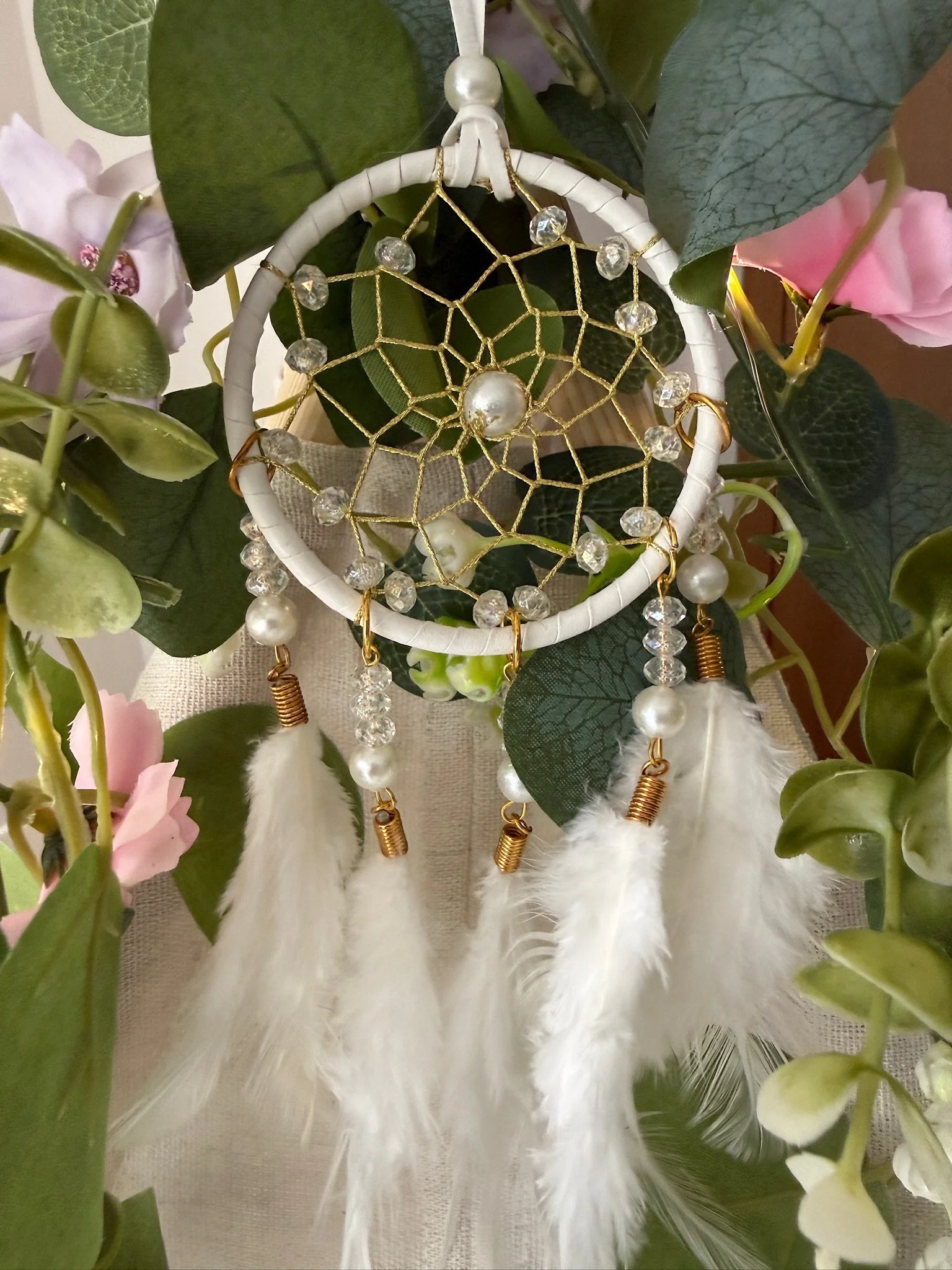 Close up detail shot of the white boho dream catcher used in the luxury teepee sleepover party hire by Sweet Dreams Events. Features pearl and crystal beads, gold tube charms, white feathers and is nestled within eucalyptus leaves and pink flowers. A