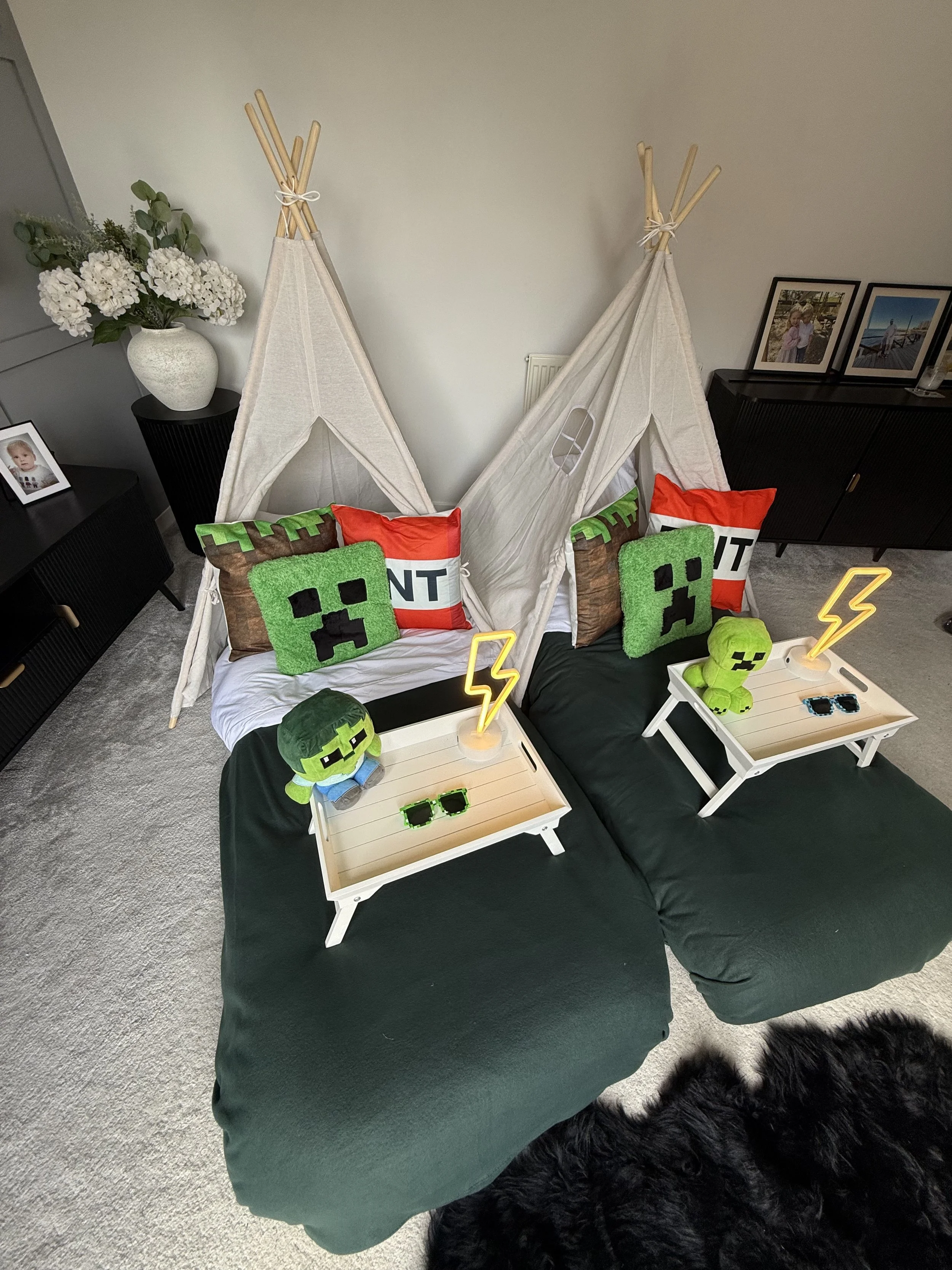 A Minecraft Sleepover teepee party setup by Sweet Dreams Events showing two cream teepees with dark green bedding, Minecraft Creeper face cushions, TNT props, Minecraft character plushies and glowing lightning bolt neon lamps on styled white breakfas