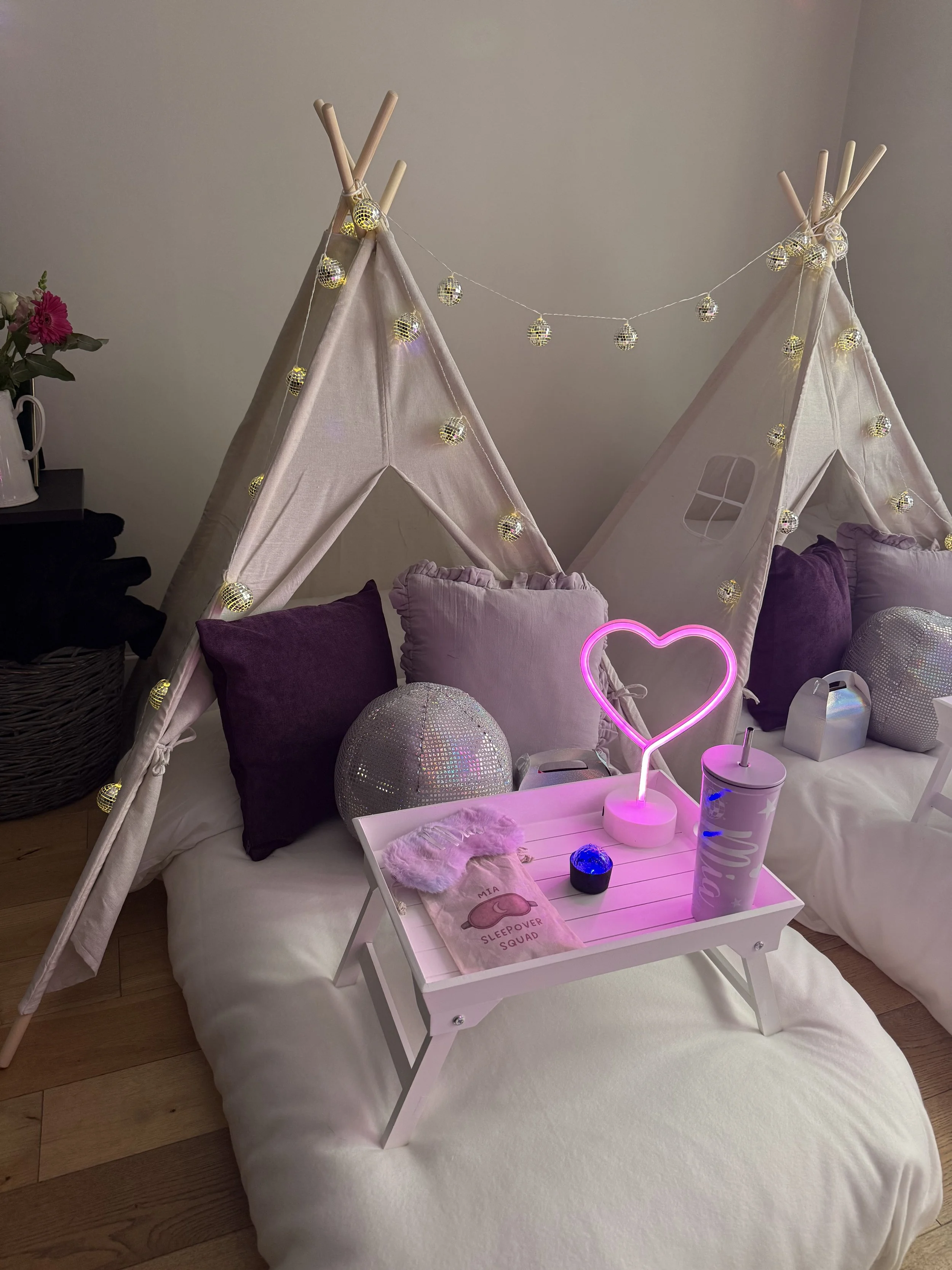 Decorative indoor setup with two small beige fabric teepees with string lights, purple and beige cushions, a white tray table with a heart-shaped pink neon light, a reusable tumbler, a small black candle, a pink travel pouch, and a plush blanket, wit