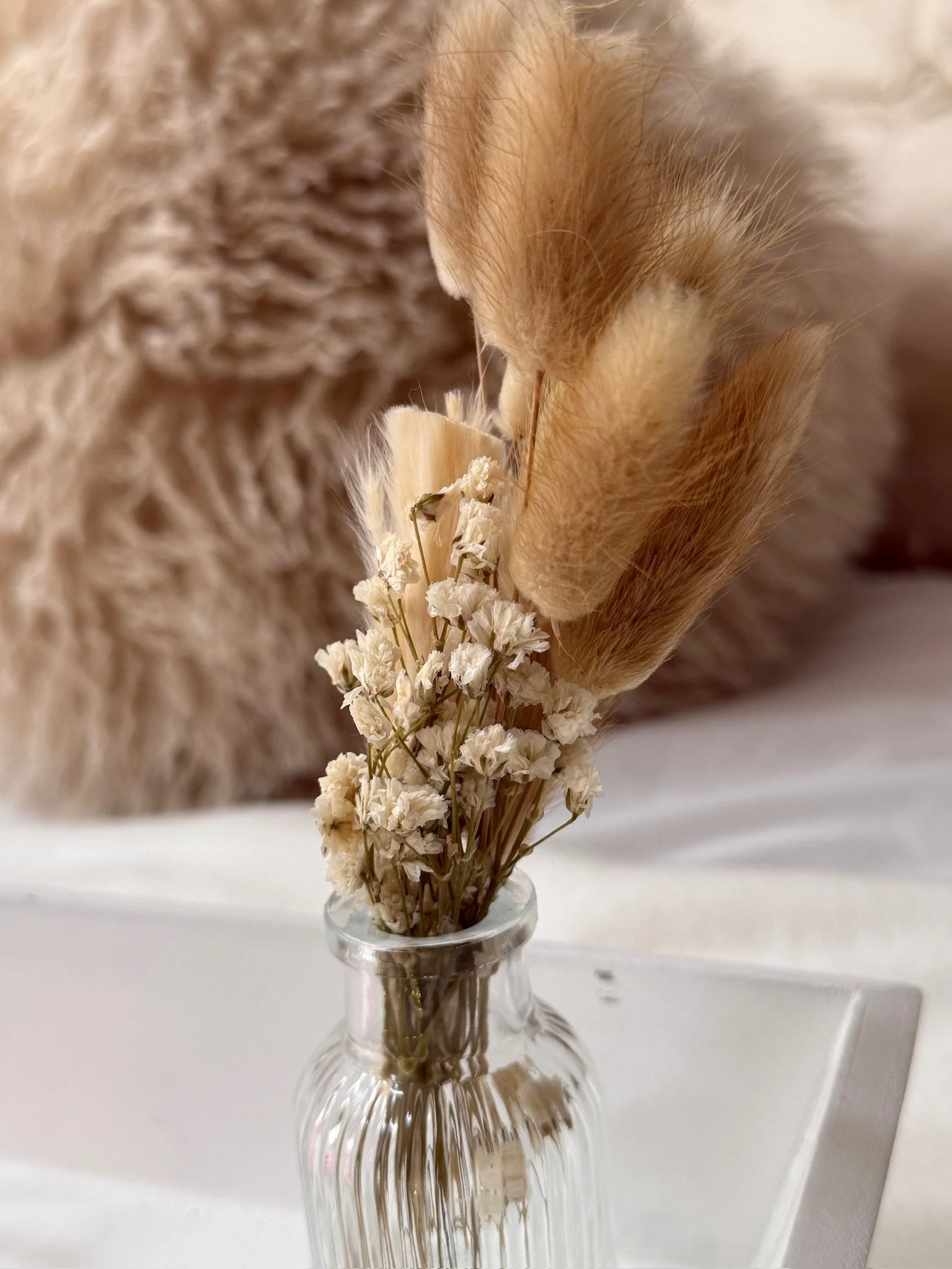 Close up detail shot of dried bunny tail grass and gypsophila in a ribbed glass vase sitting on a white styled tray, part of the boho themed teepee sleepover party hire by Sweet Dreams Events. A fluffy cushion is softly blurred in the background. Ava