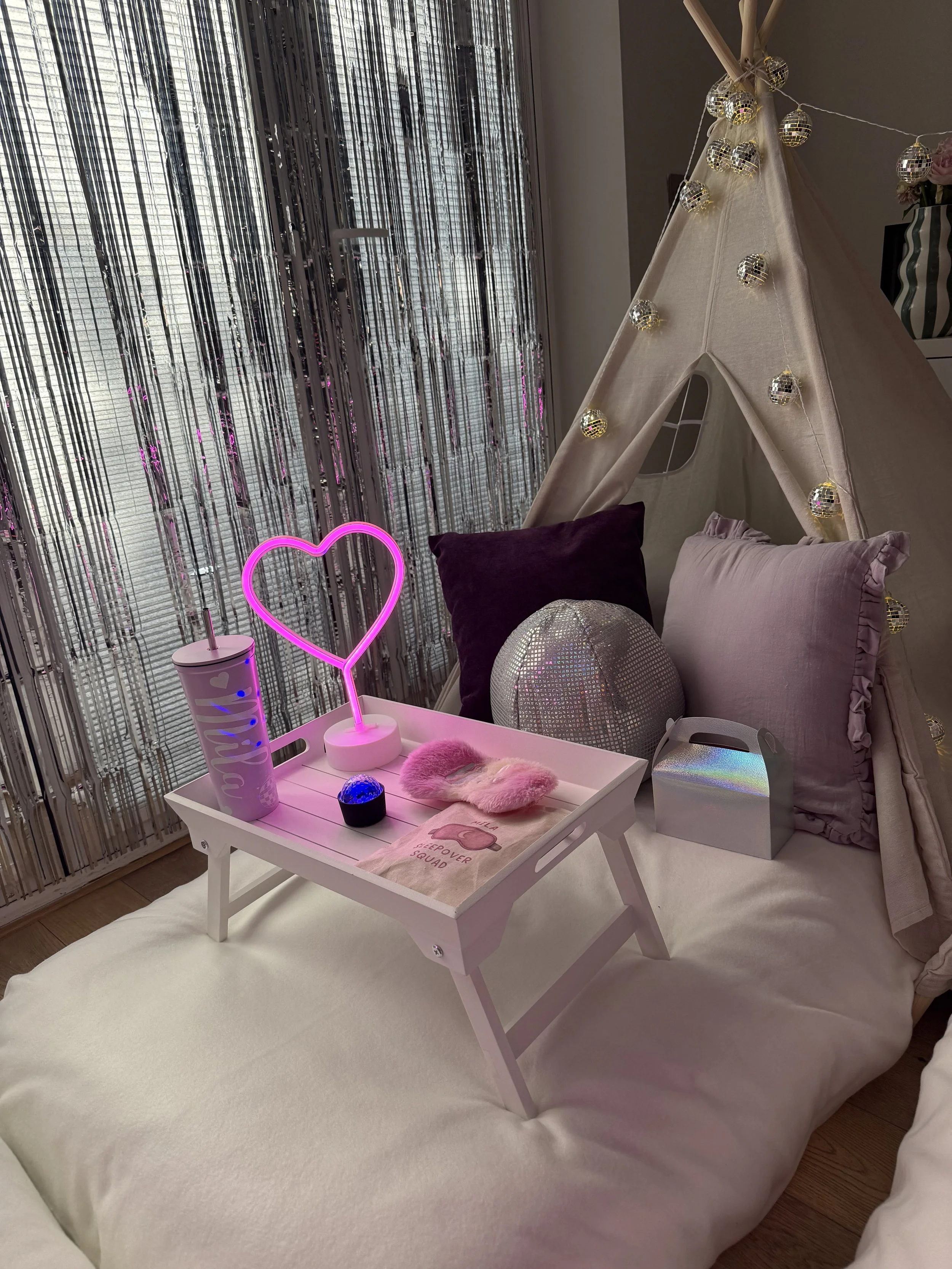 A bedroom with a white bed, a white tray on the bed holding pink fluffy slippers, a pink neck pillow, a holographic ball, and other small items. Next to the bed is a white nightstand with a pink neon heart light, a glow-in-the-dark tumbler, and a sti