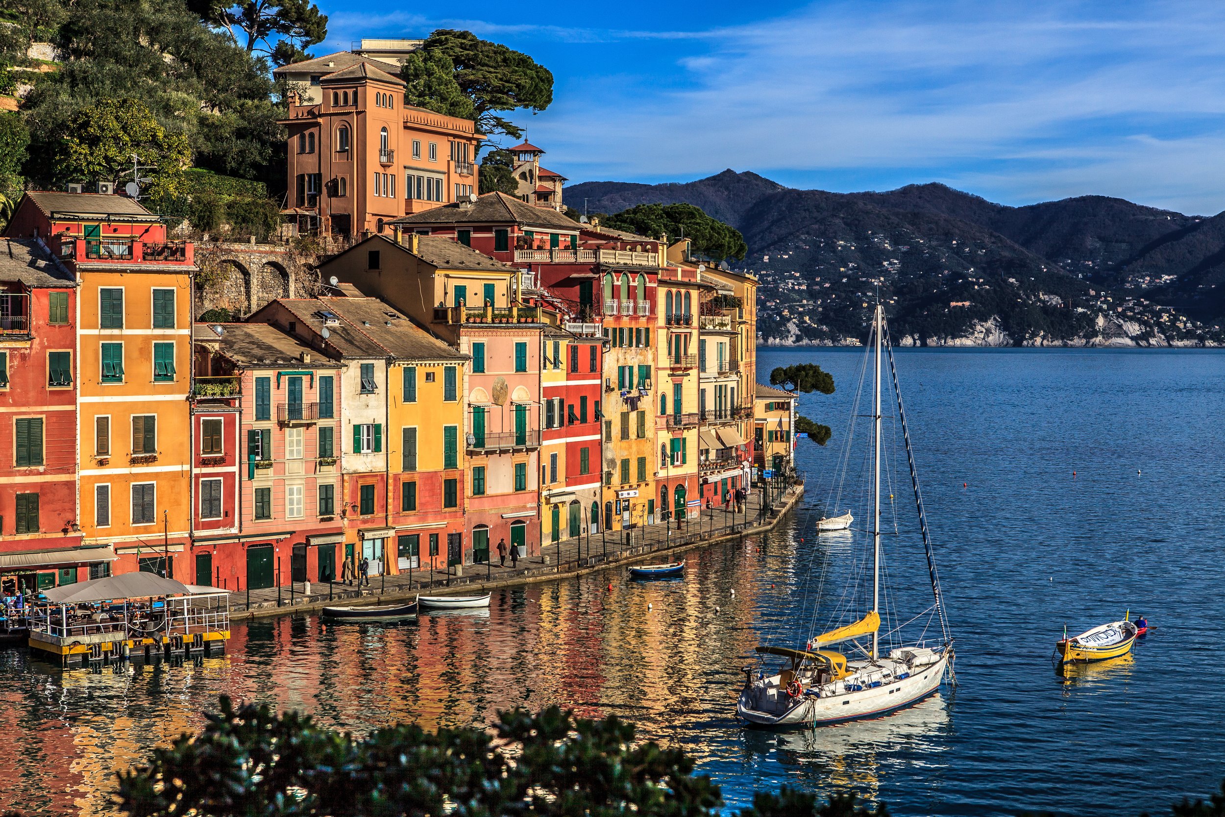 Colorful buildings along a waterfront with boats docked in the marina, hillside with trees and mountains in the background, clear blue sky.