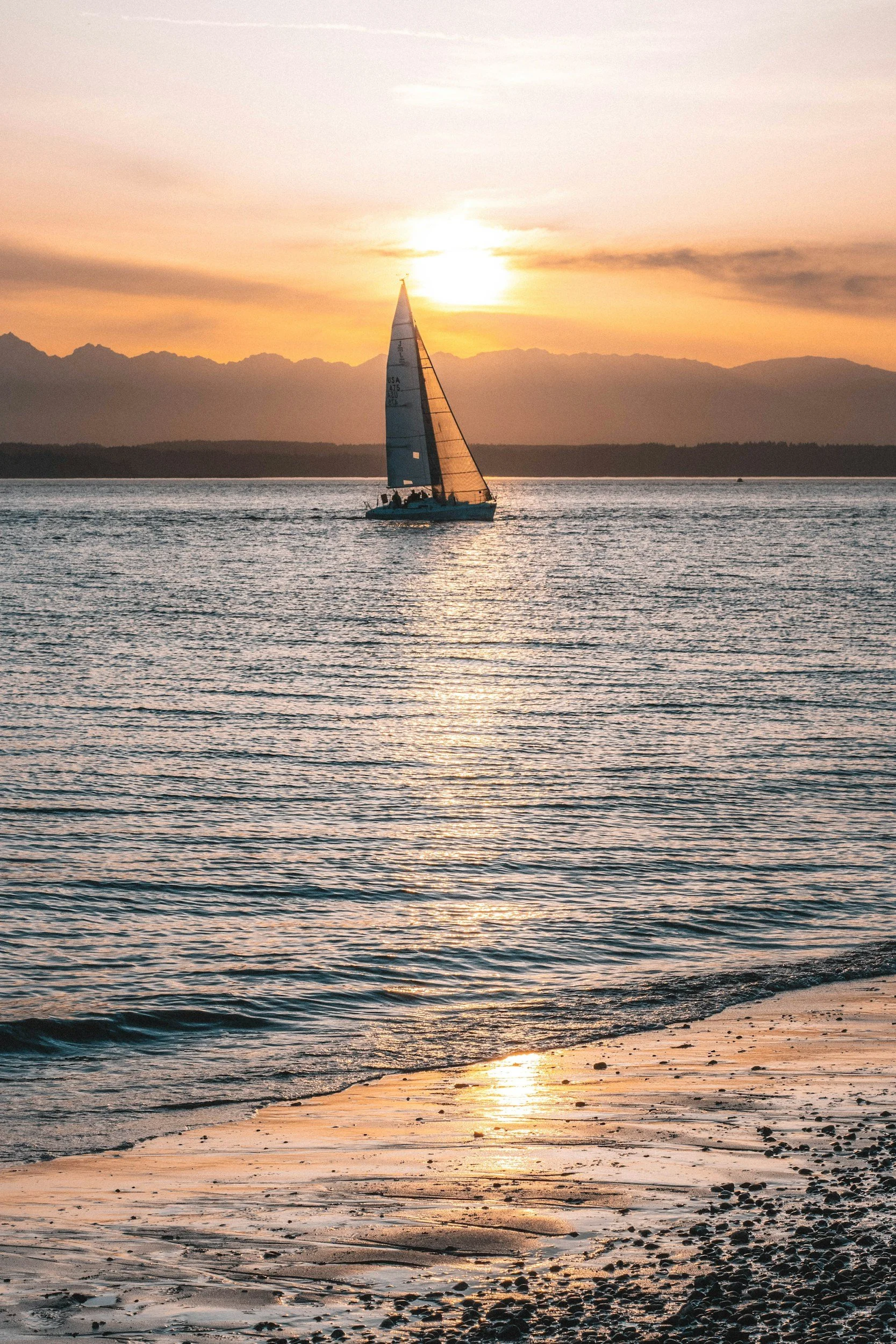 Sailing boat on calm water during sunset, mountains in the background, sandy beach in the foreground.