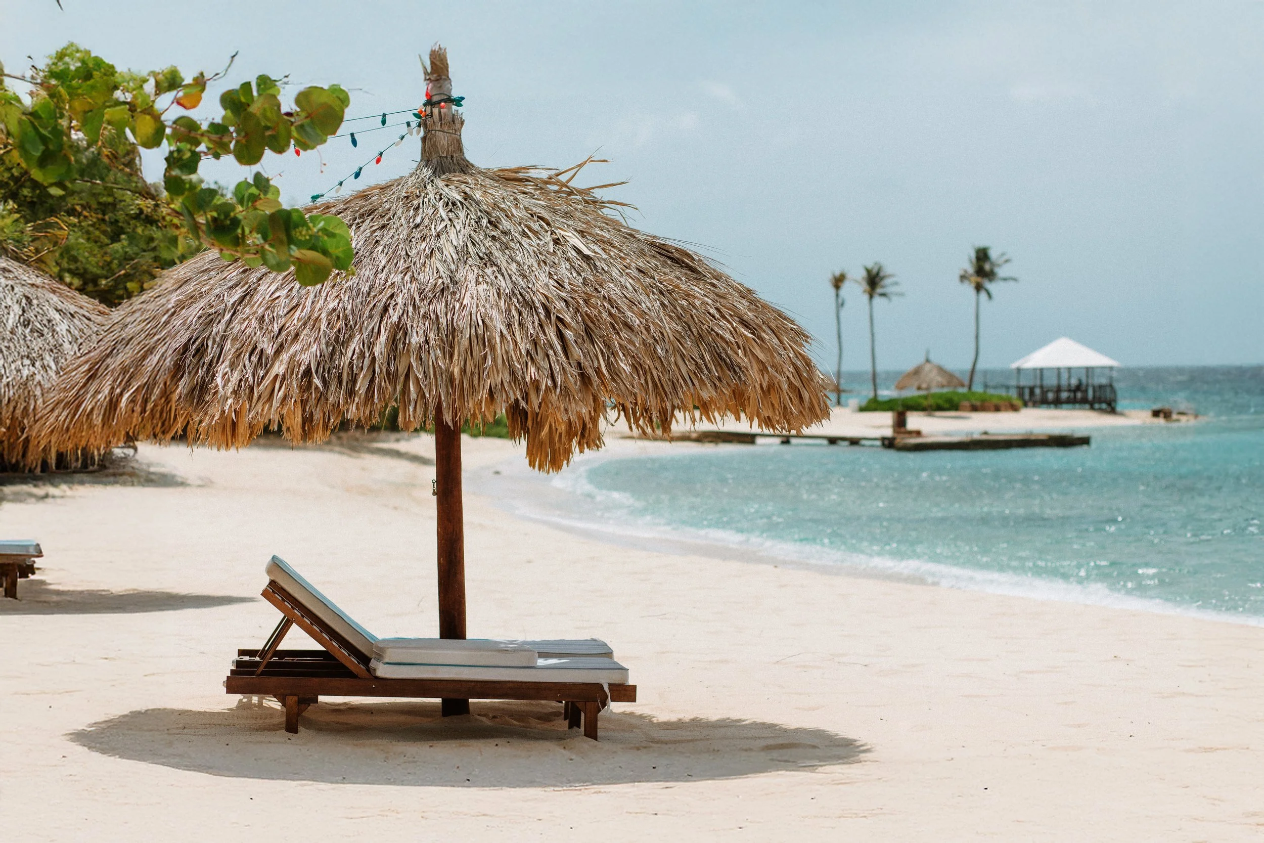 A sandy beach with a thatched-roof umbrella, a lounge chair with books, and the ocean in the background, with trees and a gazebo.