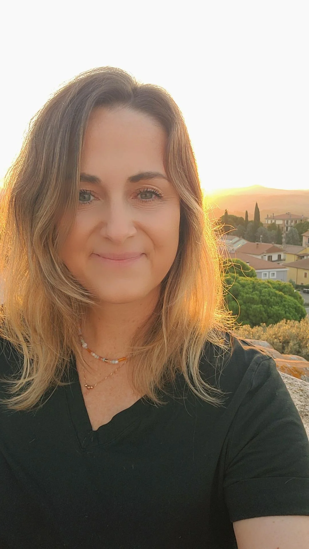 A woman with shoulder-length light brown hair stands outdoors during sunset, with a background of houses, trees, and distant hills.