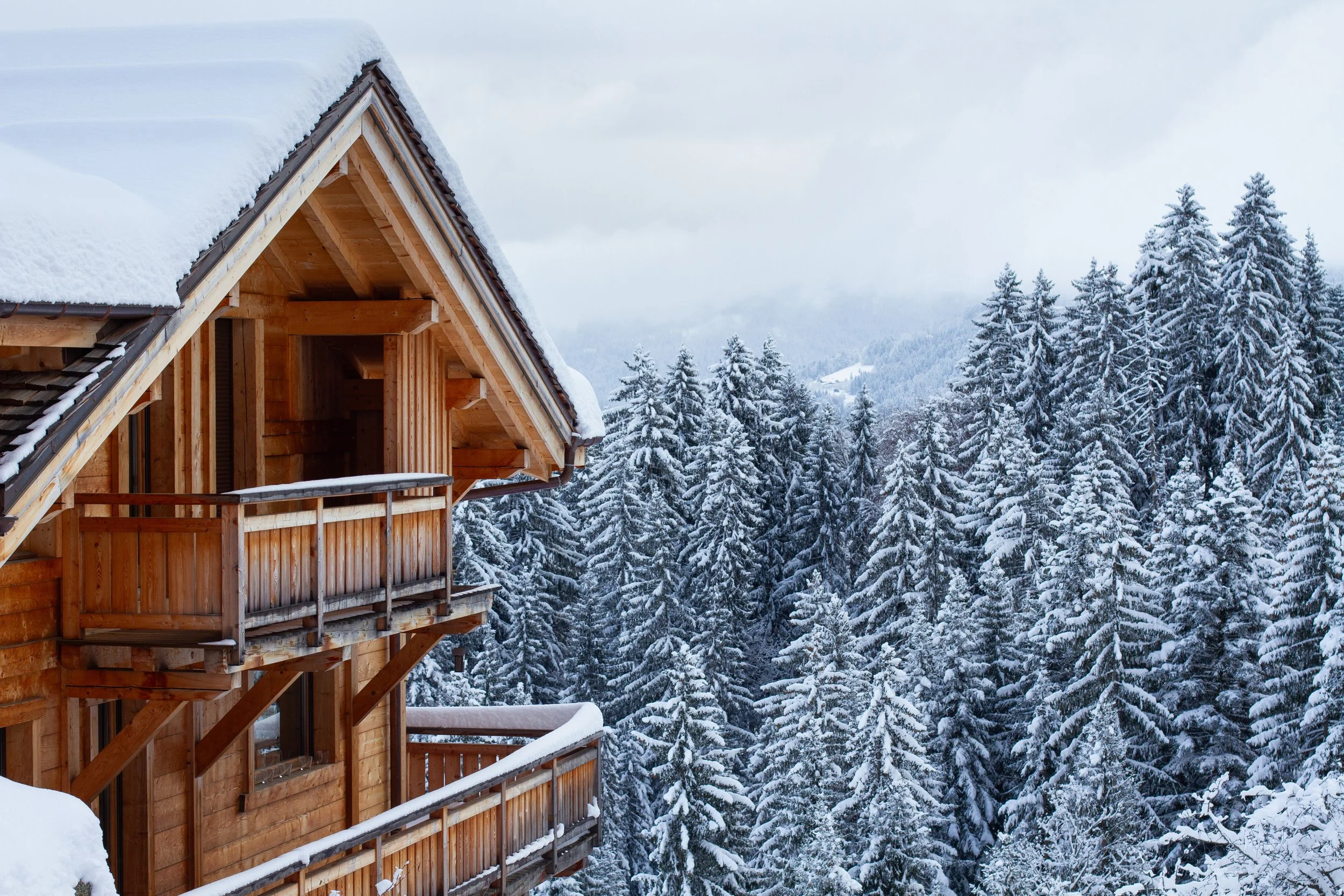 A wooden mountain ski chalet covered in snow, surrounded by snow-covered evergreen trees, with a cloudy sky in the background.