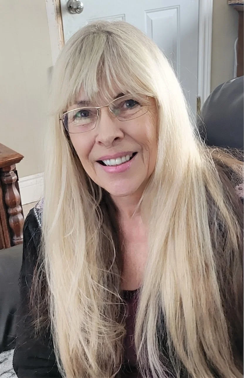 A woman with long blonde hair and glasses smiling indoors.