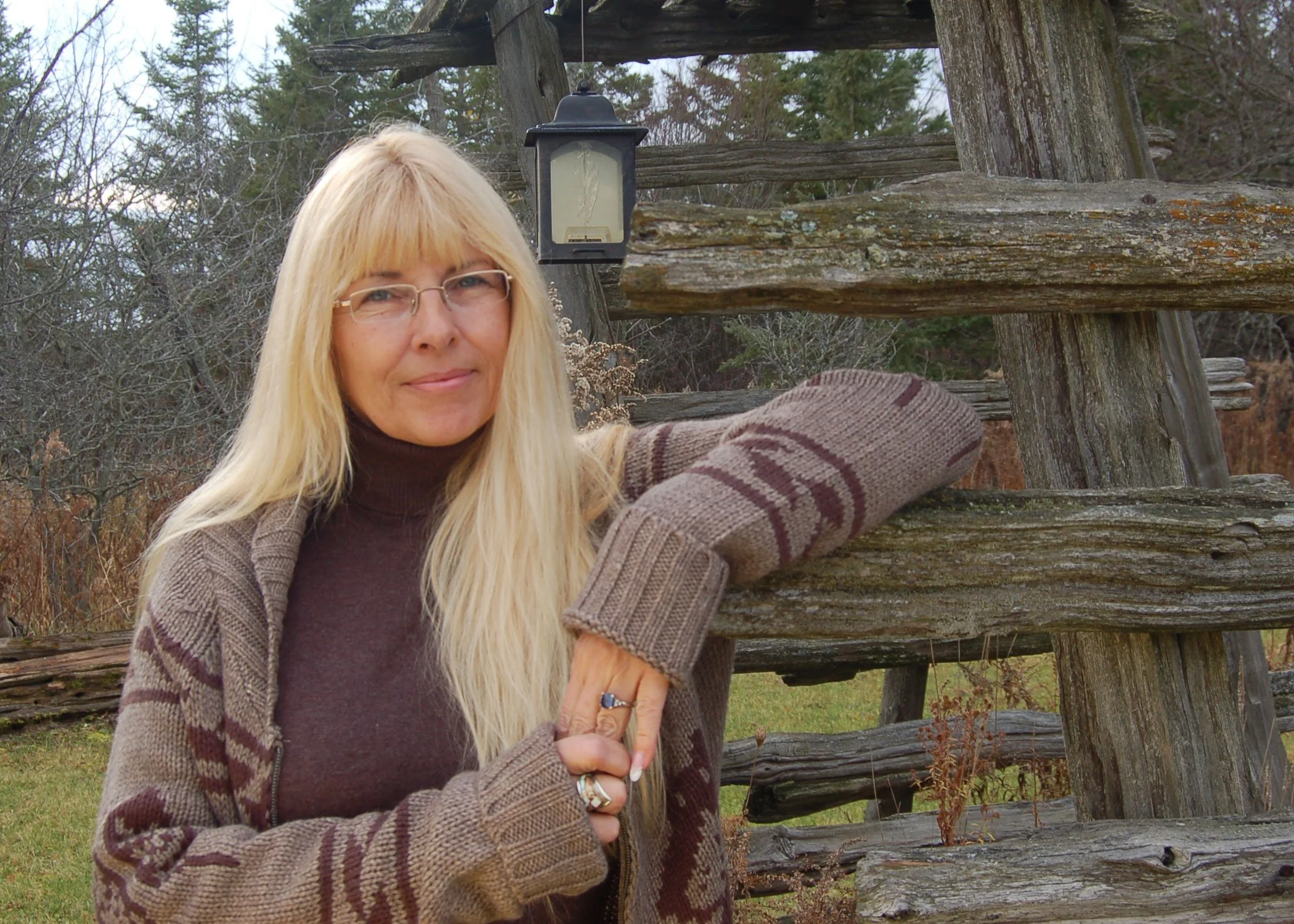 A woman with long blonde hair, glasses, wearing a brown sweater and striped cardigan, stands outside in front of a rustic wooden fence and a lantern, with trees and grass in the background.