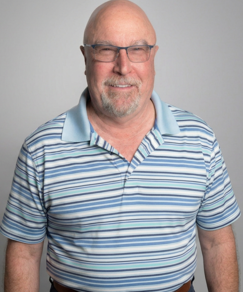 A smiling older man with glasses, wearing a striped polo shirt, standing against a plain background.