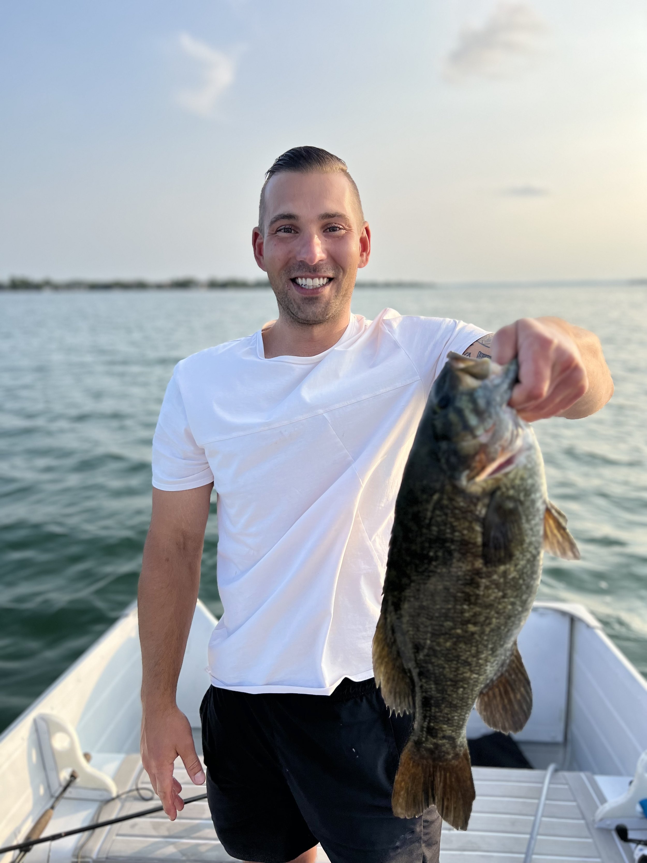 A smiling man on a boat holding a large fish in front of him, with water and a distant shoreline in the background.