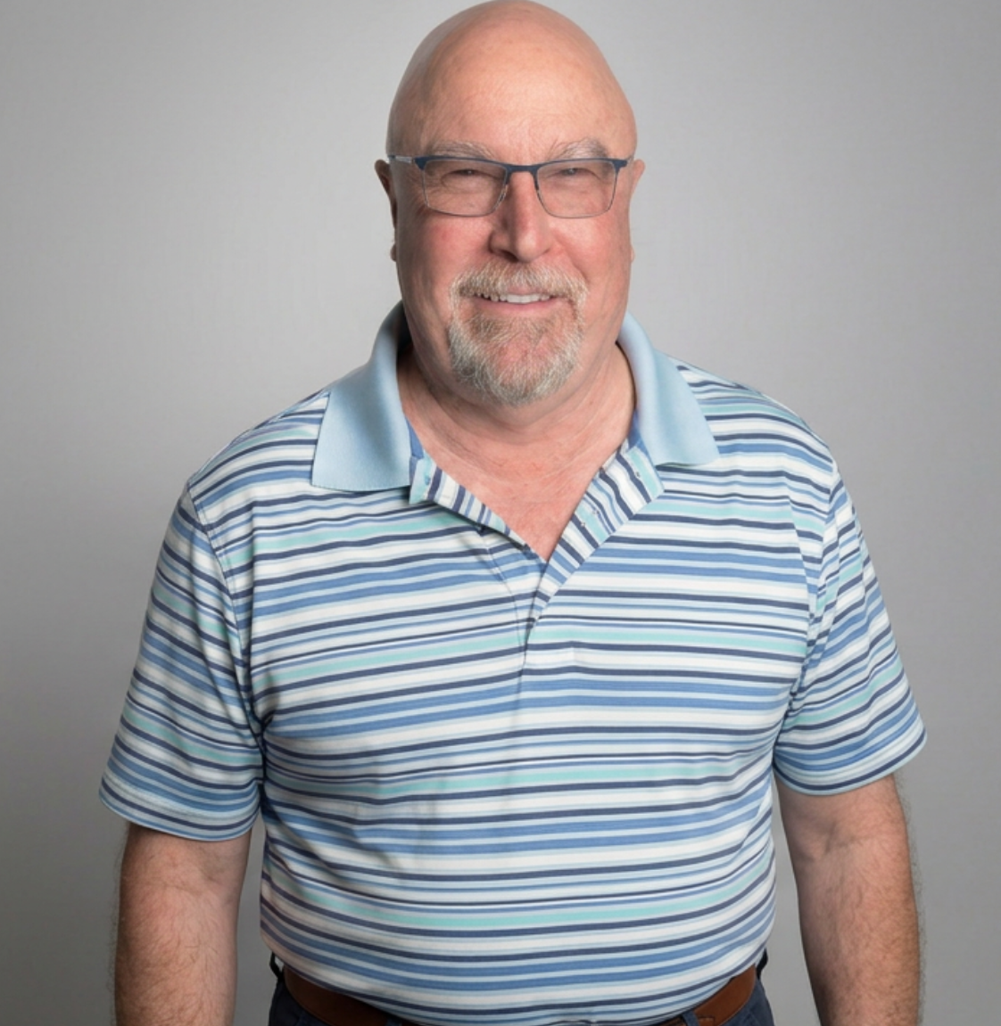 Smiling older man with glasses, wearing a striped polo shirt, standing against a plain light gray background.
