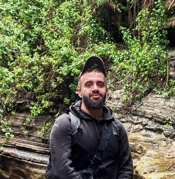 A man with a beard wearing a black jacket and a black cap, standing outdoors with green foliage and rocks in the background.