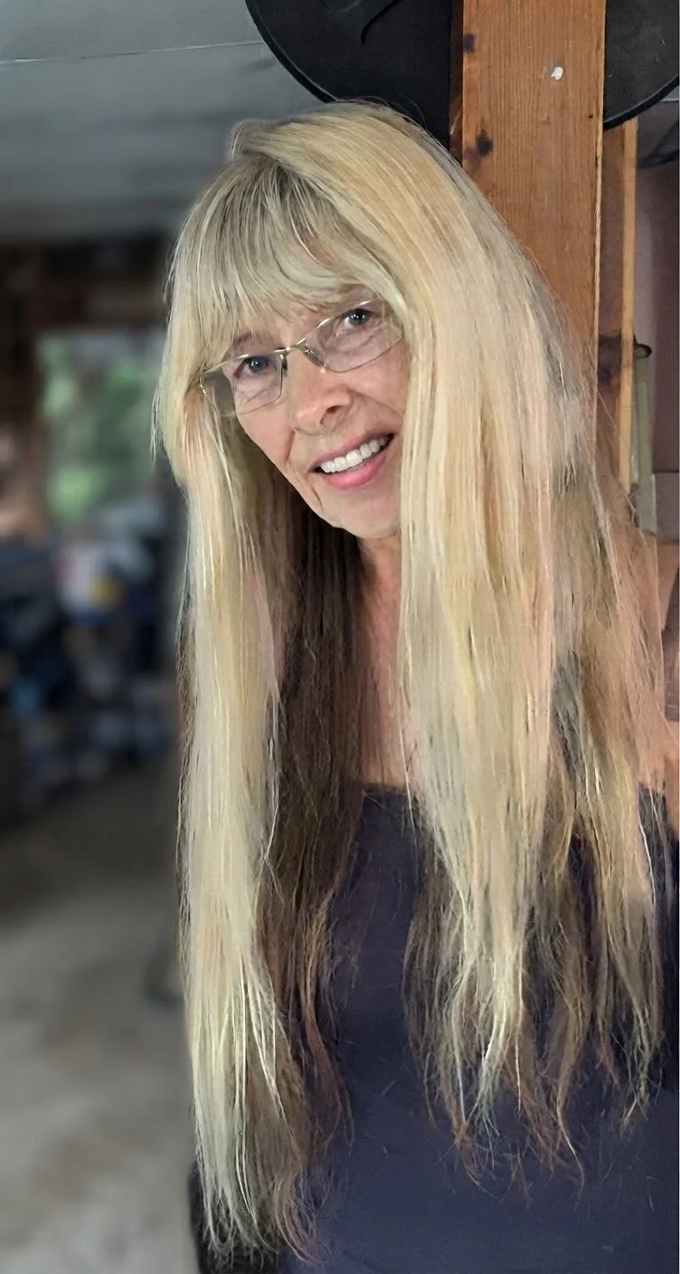 A woman with long blonde hair, glasses, and a black shirt, smiling, standing indoors with a wooden beam behind her.