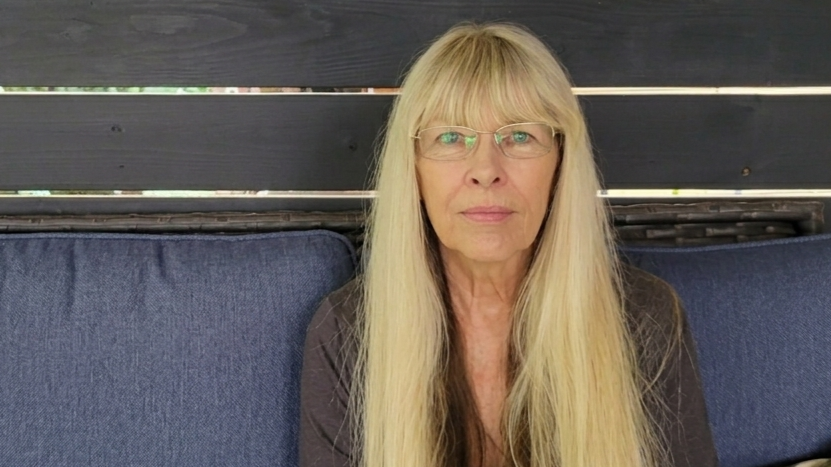 An older woman with long blonde hair, glasses, and a serious expression sitting on a blue cushioned bench in front of a dark wooden background.
