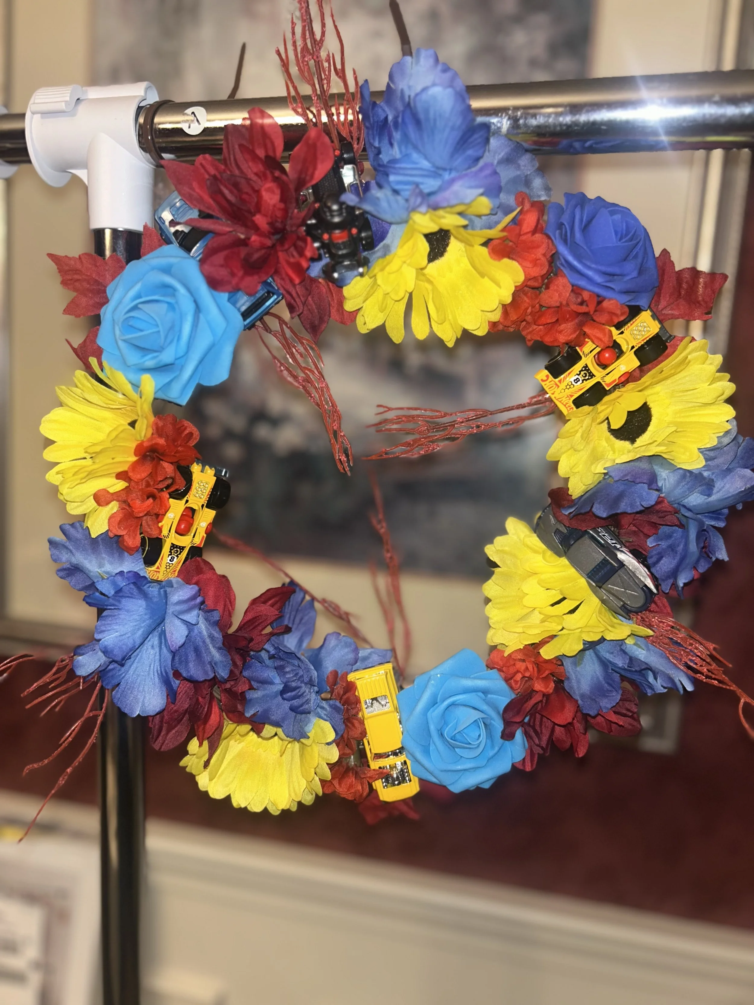 A floral wreath made of artificial blue, yellow, and red flowers, decorated with small toy yellow school buses and red leaves, hanging on a metal stand.